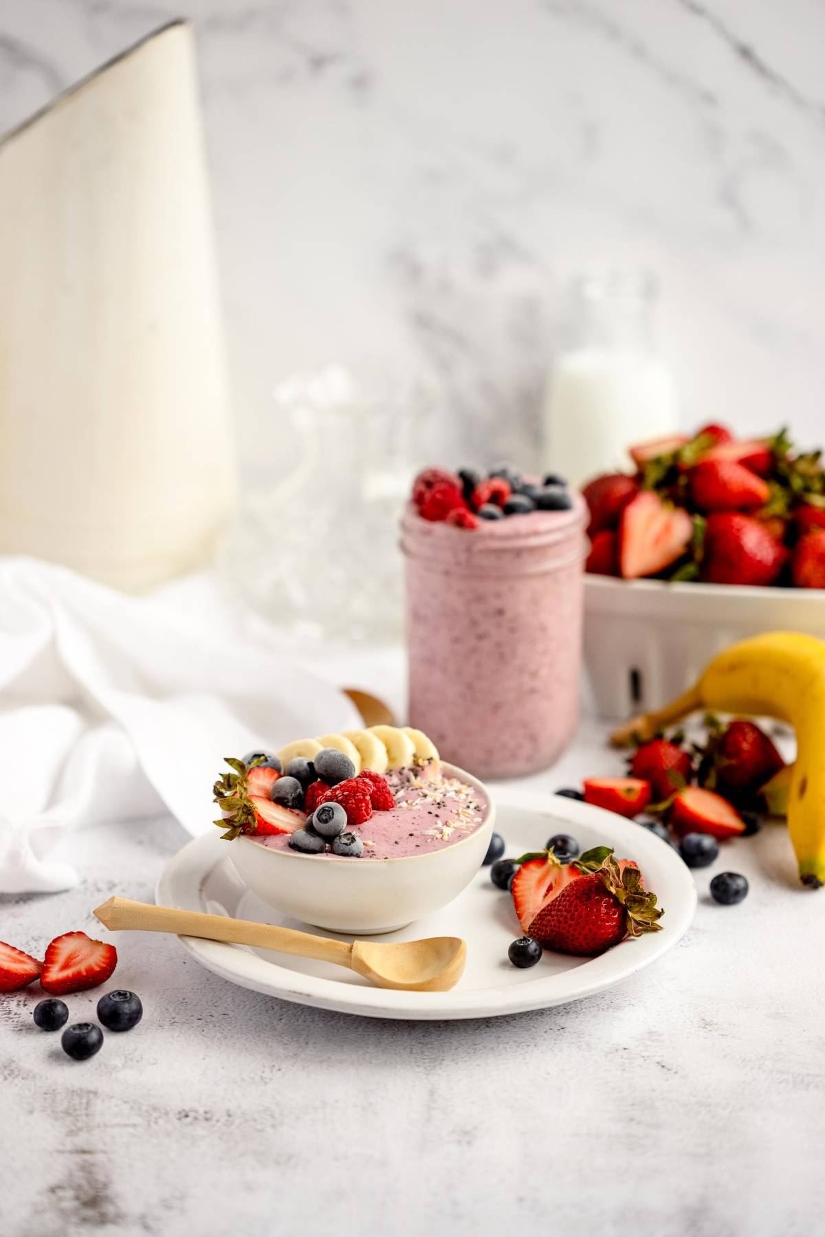 A smoothie bowl topped with berries and banana slices, surrounded by fresh fruit and a jar of smoothie, makes DIY Meal Prep easy—just use frozen smoothie bags for a quick, nutritious breakfast.