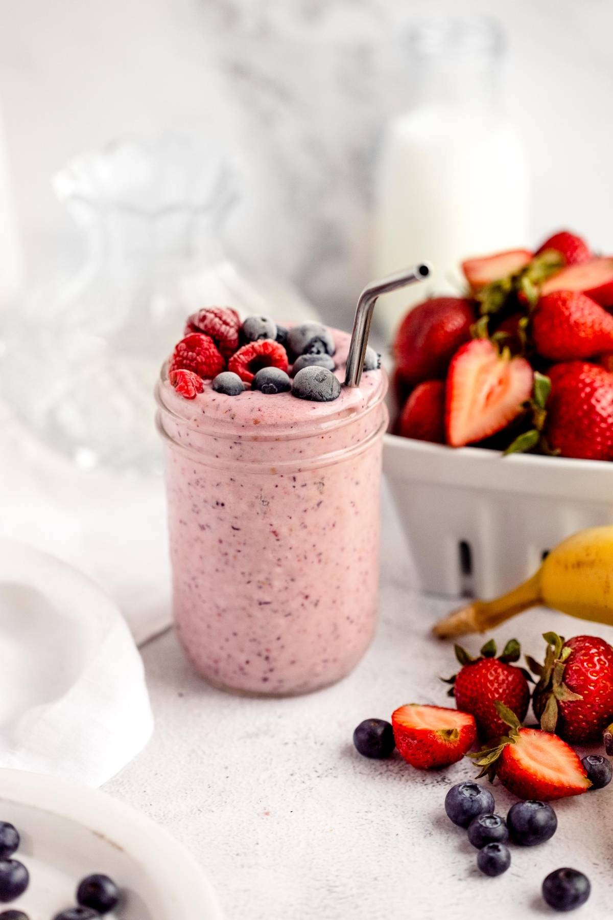 A berry smoothie in a jar with a metal straw, topped with fresh berries, sits next to strawberries and a banana—perfect for DIY Meal Prep or using a homemade smoothie pack!.