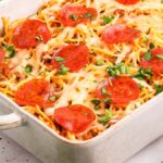 Baked spaghetti pizza casserole topped with melted cheese, pepperoni slices, and chopped parsley in a white dish.