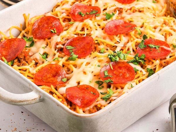 Baked spaghetti pizza casserole topped with melted cheese, pepperoni slices, and chopped parsley in a white dish.