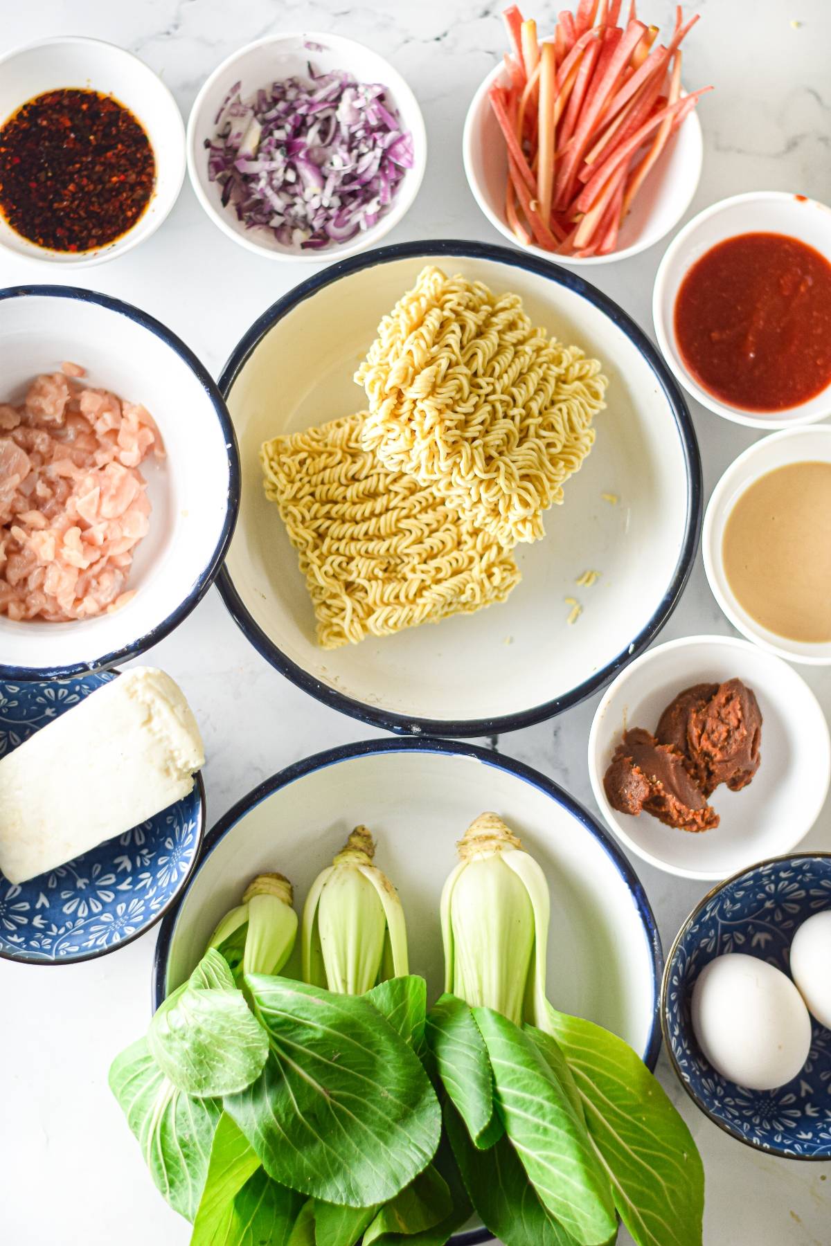 Bowls with raw noodles, bok choy, chicken, sauces, egg, daikon, chopped onions, and shredded carrots on a white surface—perfect for trying your own spicy chicken miso ramen recipe at home.