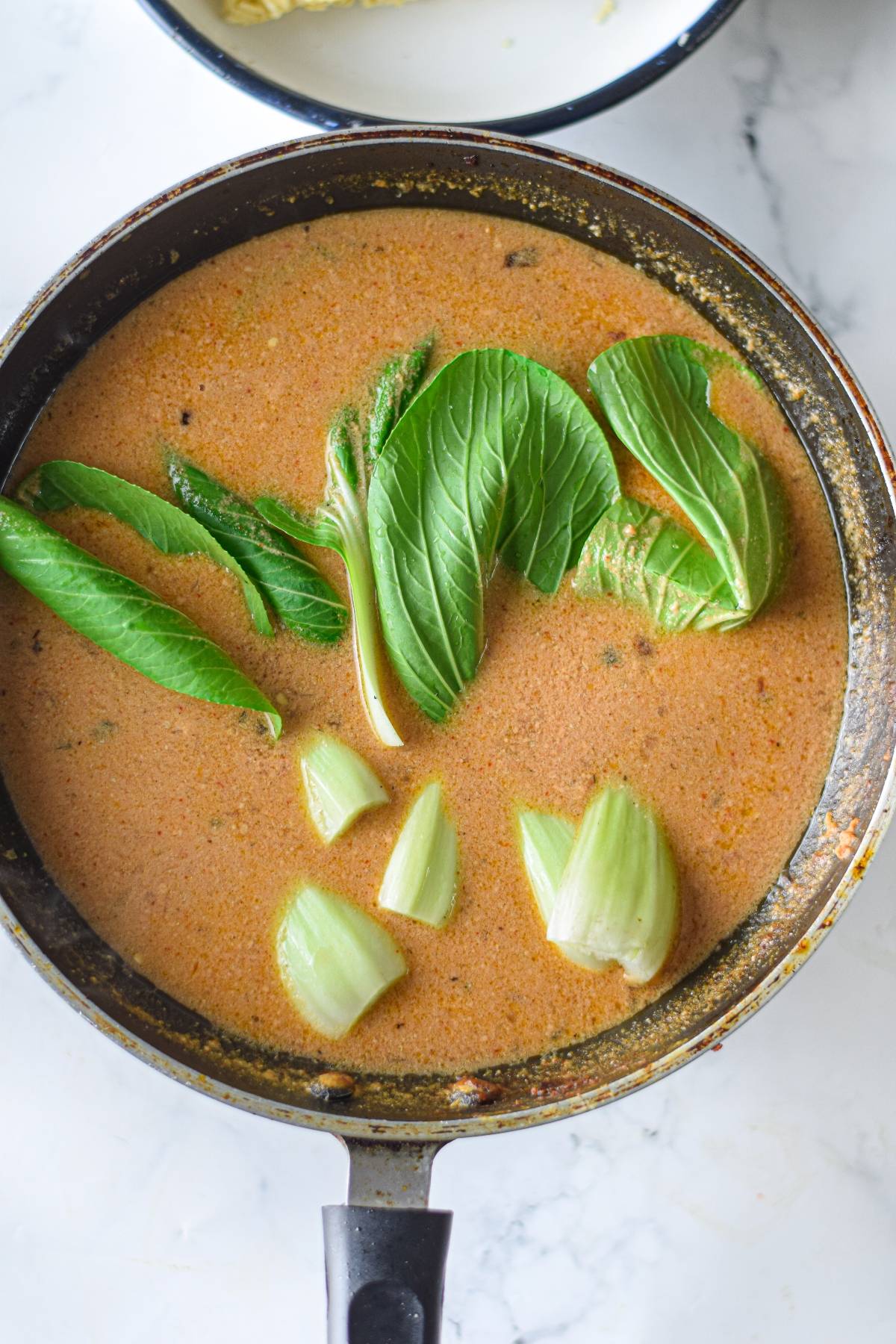 A pot of reddish Spicy Chicken Miso Ramen with bok choy and green vegetables sits on a white marble surface.