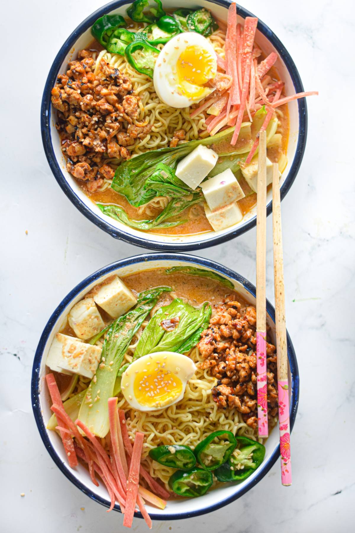 Two bowls of Spicy Chicken Miso Ramen with egg, tofu, greens, sliced meat, jalapeños, and chopsticks on a white surface.