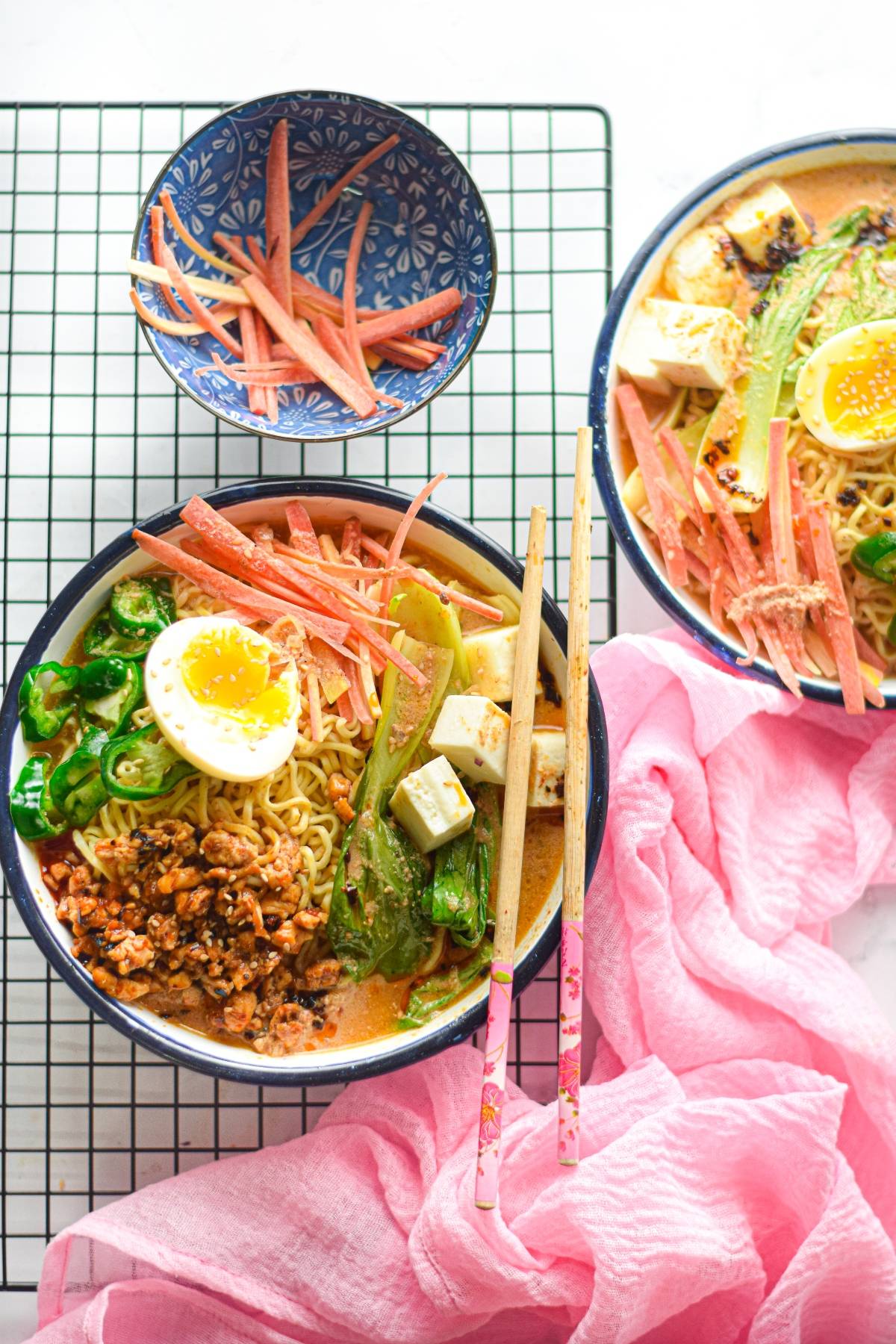 Two bowls of ramen with egg, tofu, veggies, and crab sticks on a cooling rack, pink cloth beside them—a mouthwatering ramen recipe perfect for cozy nights in.