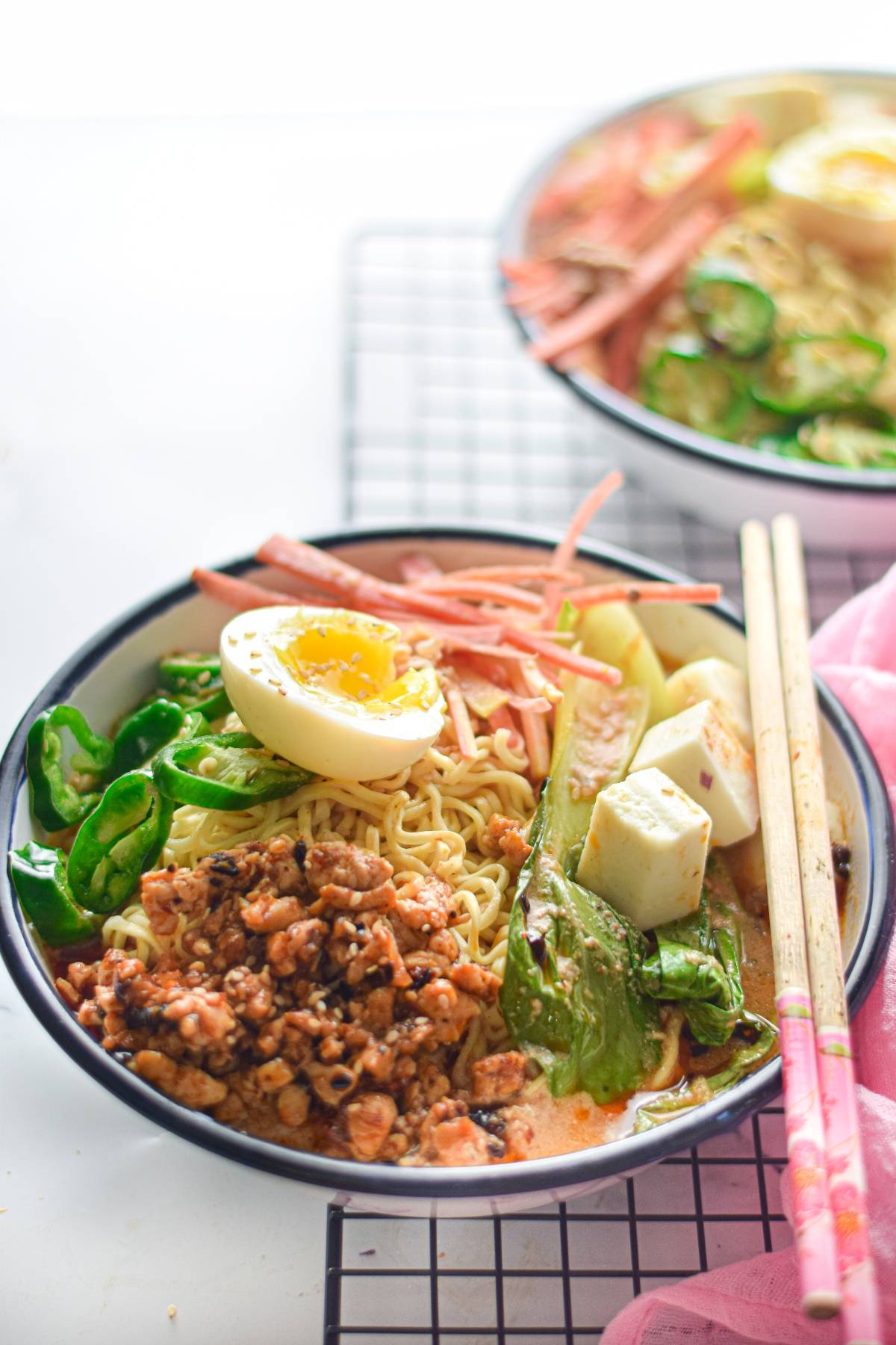 A bowl of Spicy Chicken Miso Ramen with egg, ground meat, tofu, greens, and chopsticks sits beside another bowl in the background—perfect for anyone seeking a comforting ramen recipe.