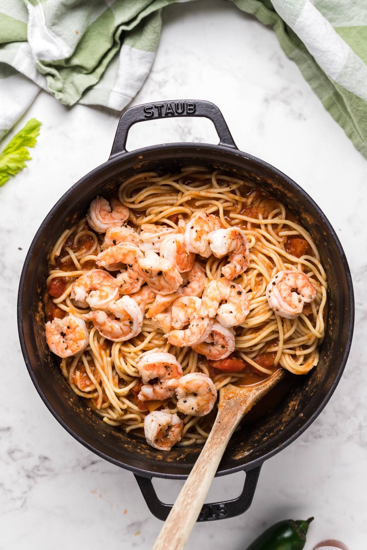 Shrimp pasta with Bloody Mary vodka sauce in a black pot, with a wooden spoon and green linens nearby on a marble surface.
