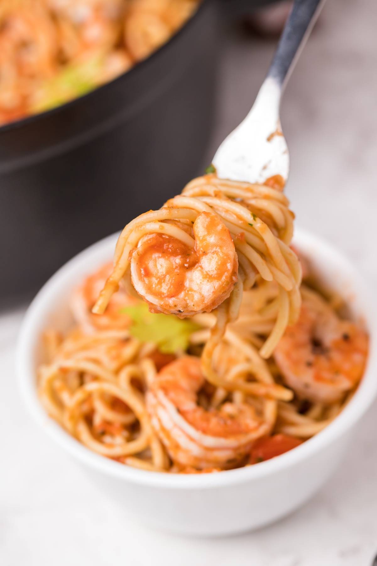 A fork with spicy shrimp pasta above a white bowl filled with shrimp and spaghetti tossed in a zesty Bloody Mary vodka sauce.