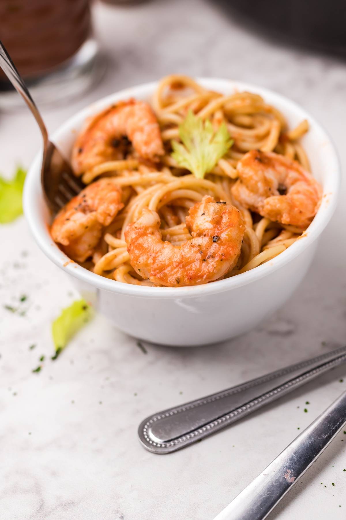 A bowl of spicy shrimp pasta with Bloody Mary vodka sauce sits with a fork and knife on a white marble surface.