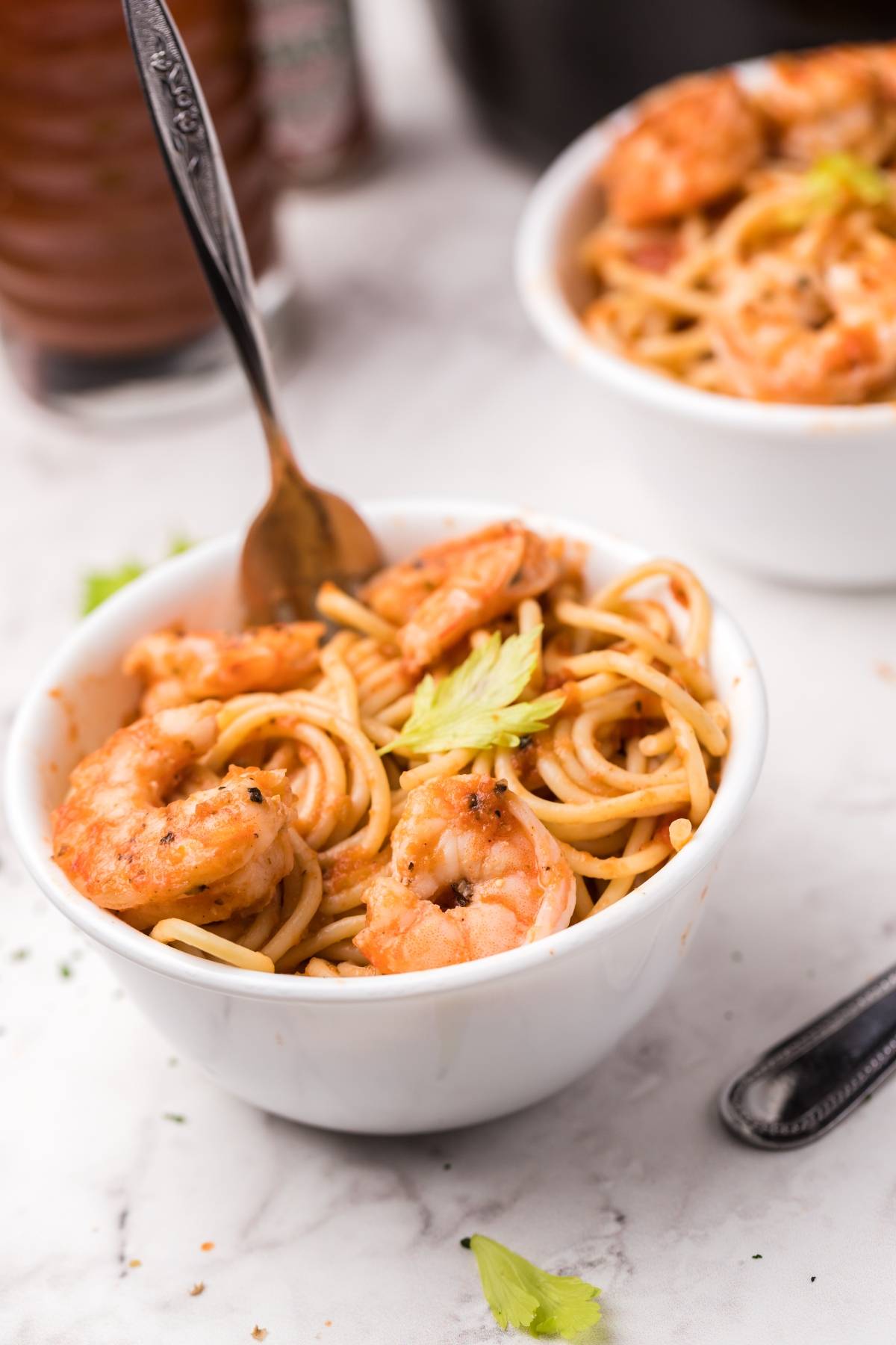 A white bowl filled with spicy shrimp pasta and spaghetti in a rich Bloody Mary vodka sauce, garnished with a celery leaf and served with a fork.