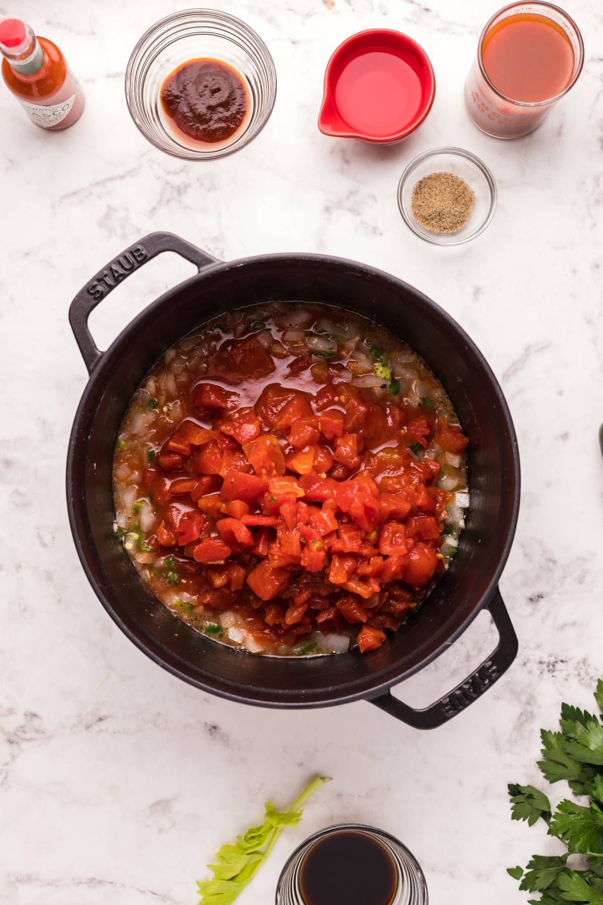 Overhead view of a pot with diced tomatoes, onions, and spices for Bloody Mary vodka sauce, surrounded by ingredients on a marble counter.