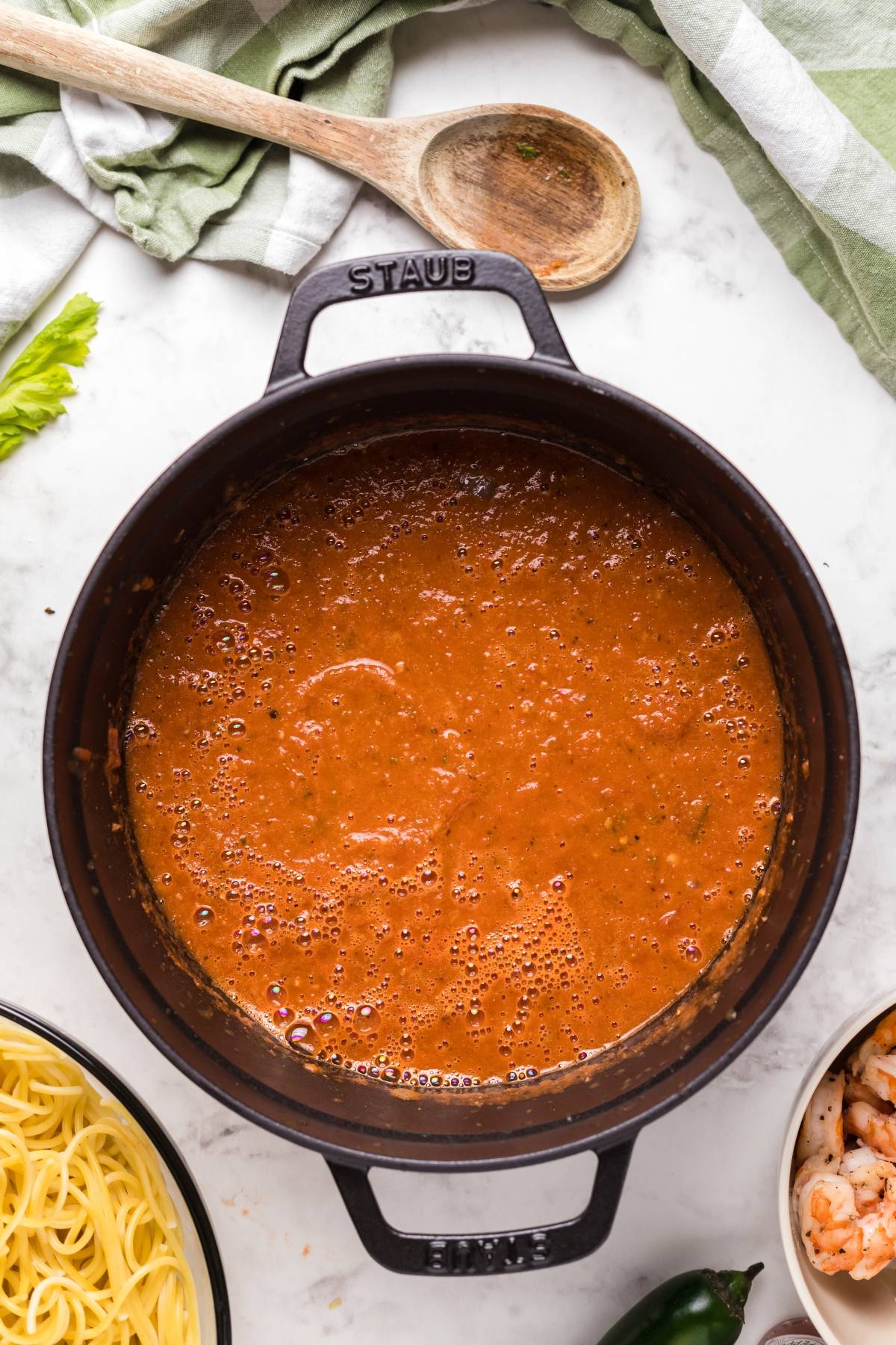 Black pot filled with rich Bloody Mary vodka sauce on a marble counter, surrounded by pasta, spicy shrimp, and a wooden spoon—perfect for making spicy shrimp pasta.