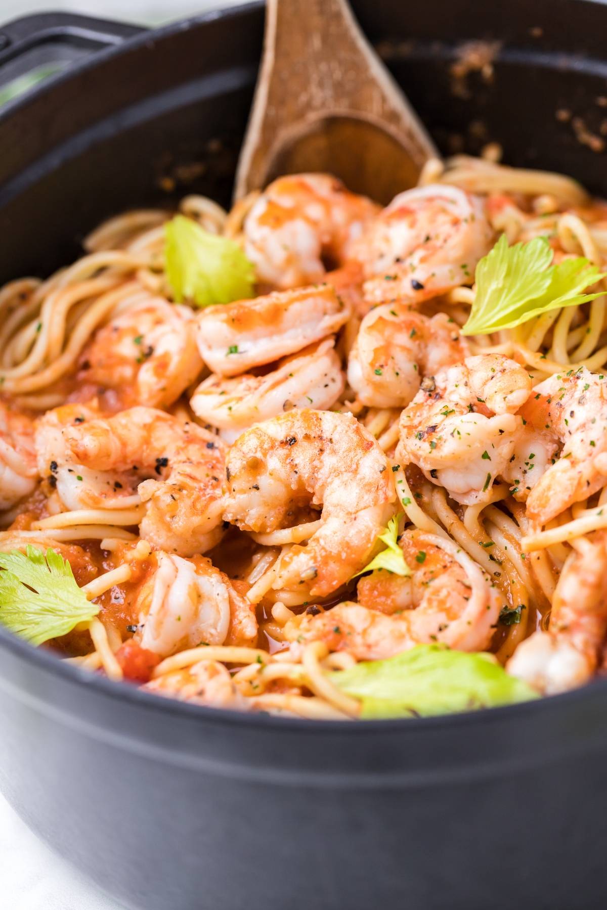 Shrimp and spaghetti in a spicy Bloody Mary vodka sauce, garnished with herbs and celery leaves, served in a black pot with a wooden spoon.