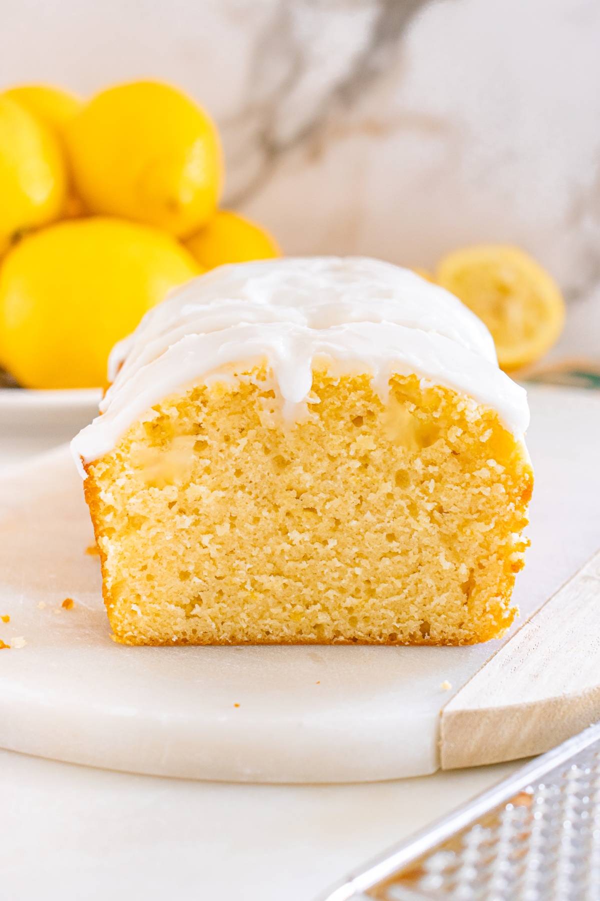 A sliced lemon loaf cake with white icing, shown on a marble board with fresh lemons in the background.