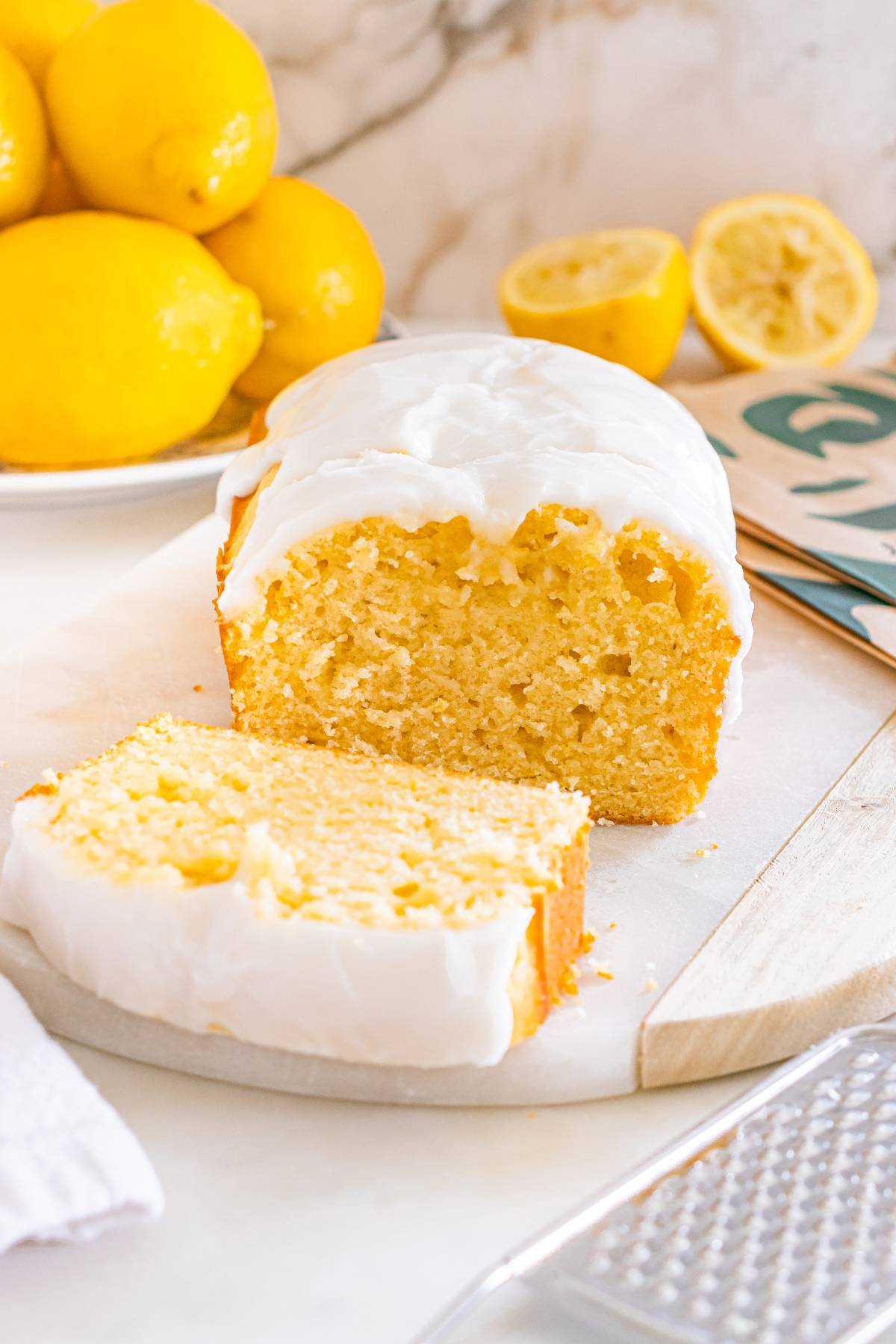 Lemon loaf cake with white icing, sliced on a cutting board, fresh lemons in the background.