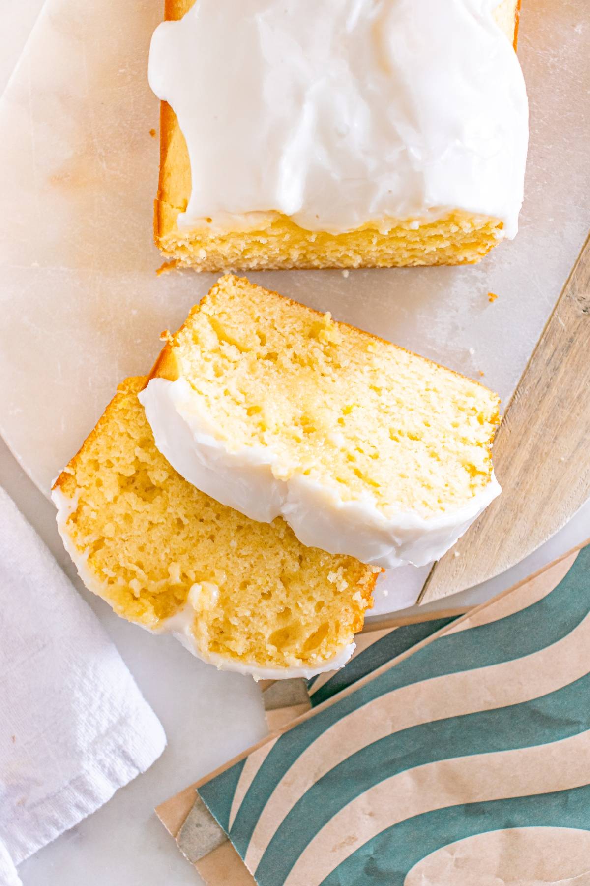 Sliced lemon loaf cake with white icing on a marble board, next to a green-striped napkin.