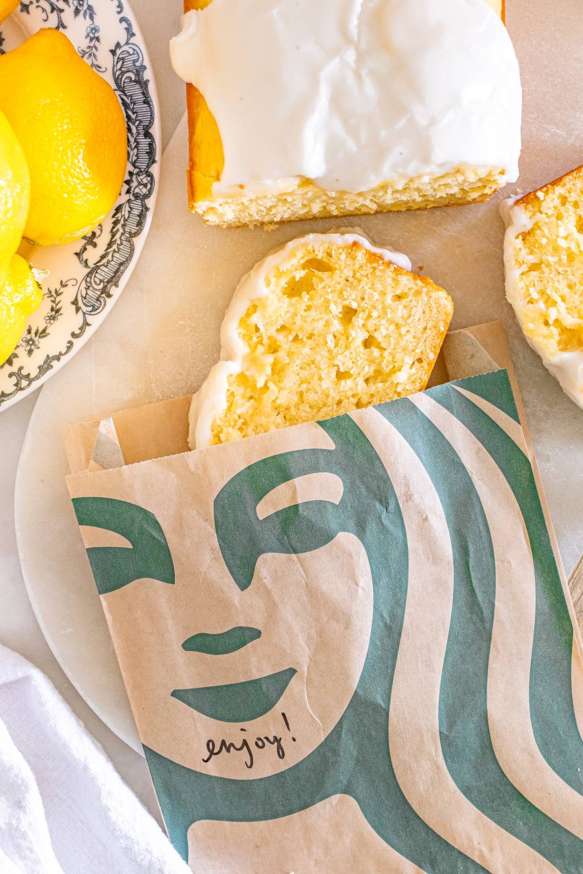 A slice of iced lemon loaf in a Starbucks bag beside the rest of the loaf and fresh lemons on a plate.