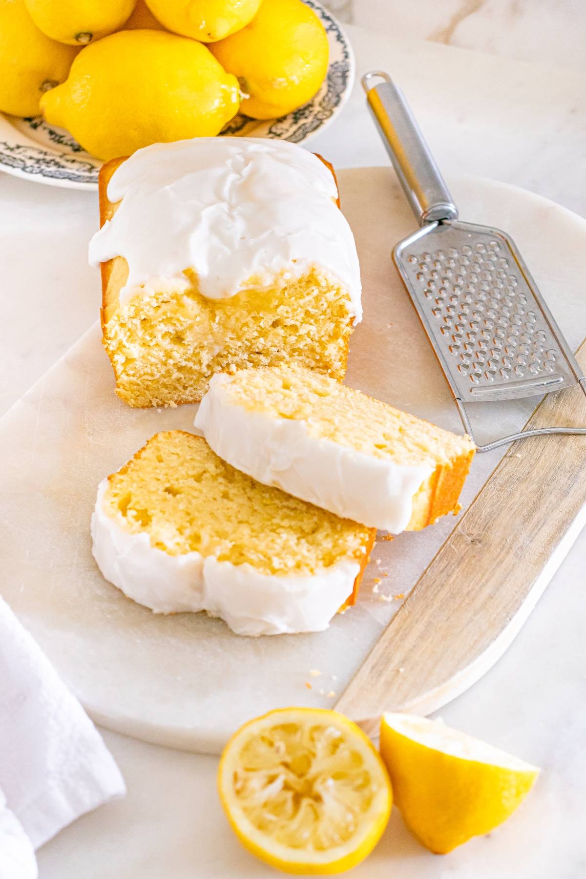 A lemon loaf cake with white icing, two slices cut, surrounded by fresh lemons and a grater on a marble board.
