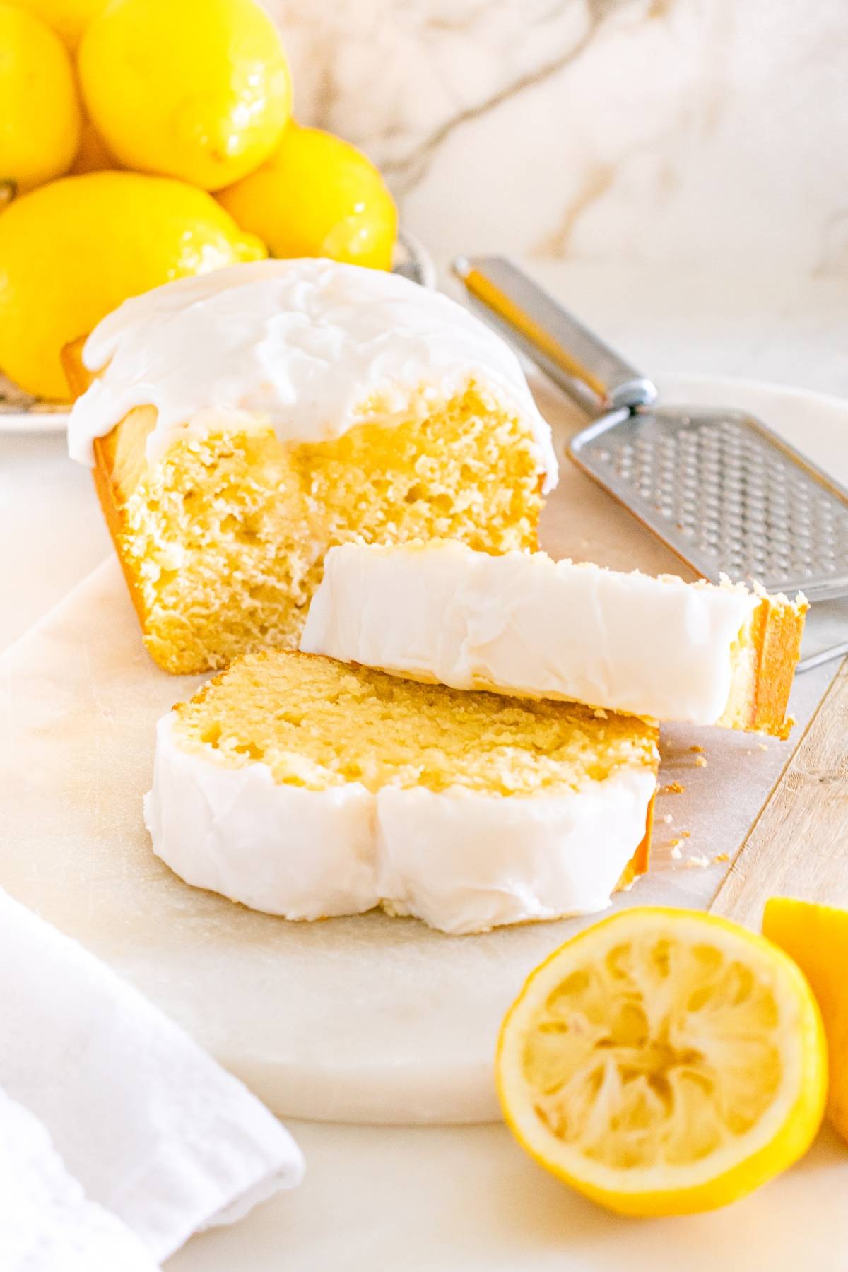 A lemon loaf cake with white icing, sliced, surrounded by fresh lemons and a zester on a marble surface.