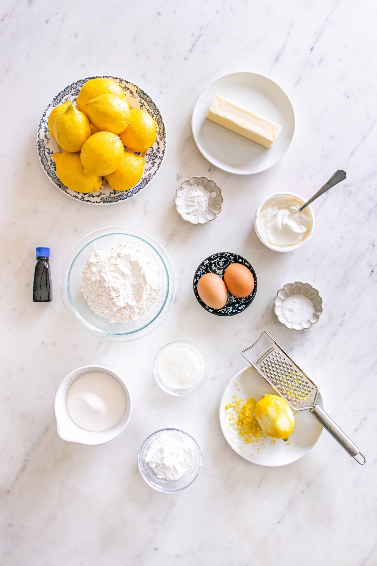 Baking ingredients, including lemons, eggs, flour, sugar, butter, and vanilla, arranged on a white countertop.