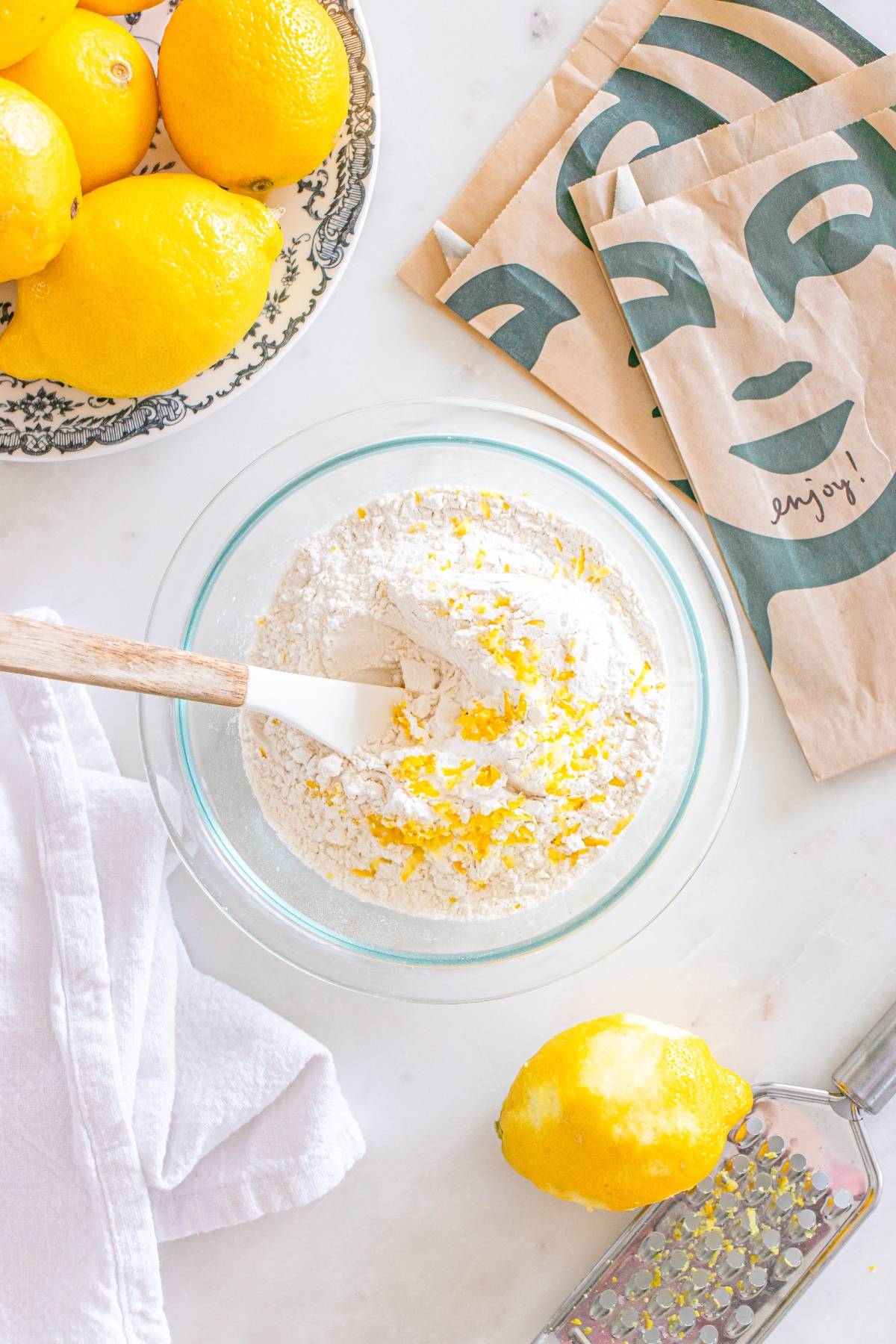A bowl of flour and lemon zest with a spatula, surrounded by lemons, napkins, and a white cloth.