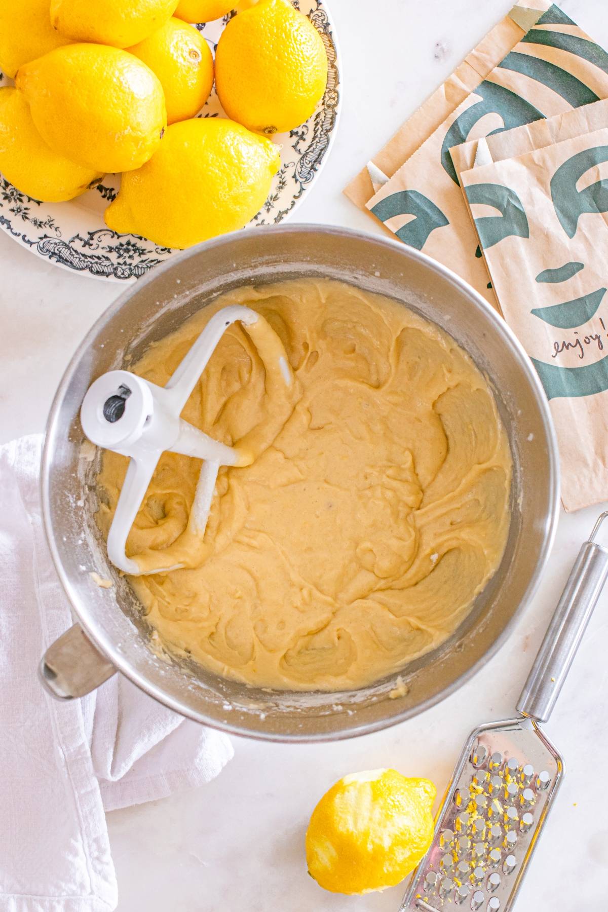 A mixing bowl with lemon cake batter, surrounded by lemons, a grater, and paper bags on a countertop.