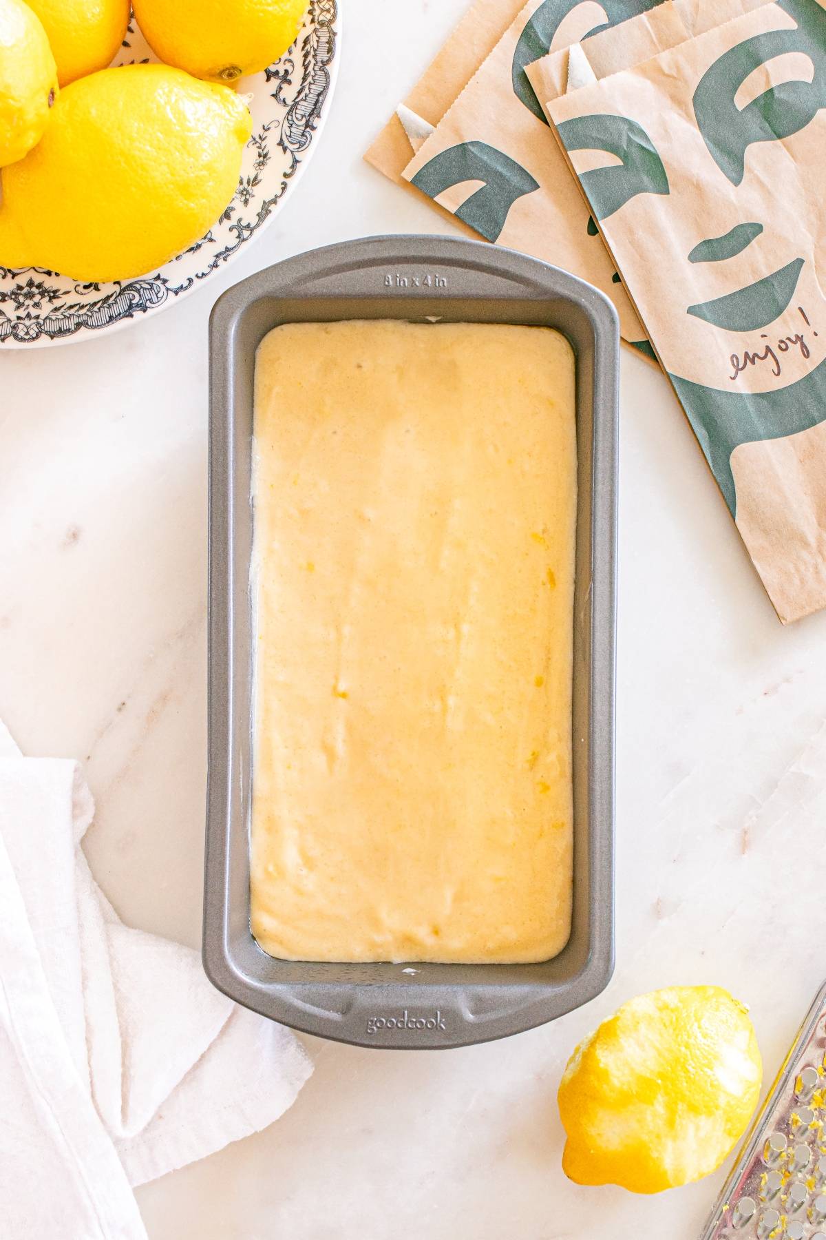 A loaf pan filled with lemon cake batter sits on a countertop next to fresh lemons and a paper napkin.