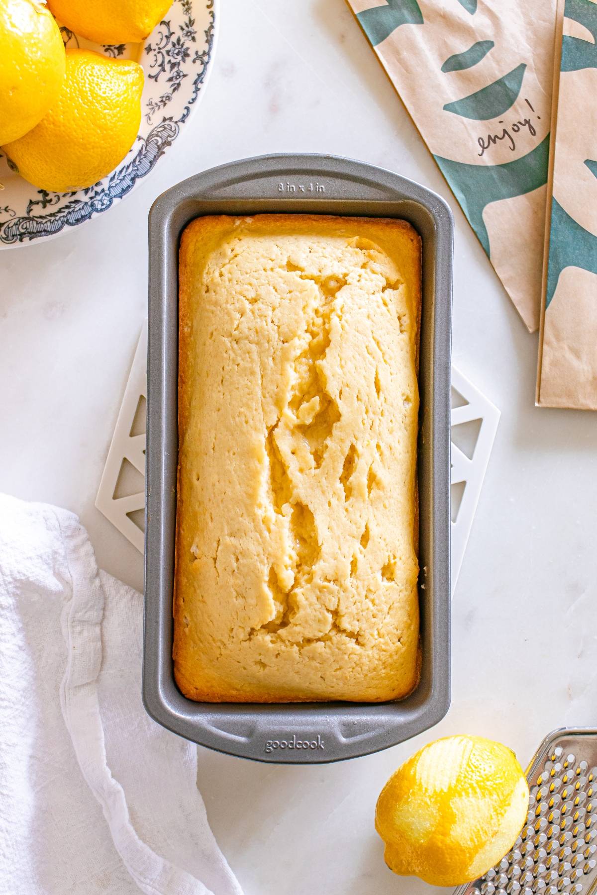 A loaf of lemon bread in a baking pan on a white surface, surrounded by lemons and a kitchen towel.