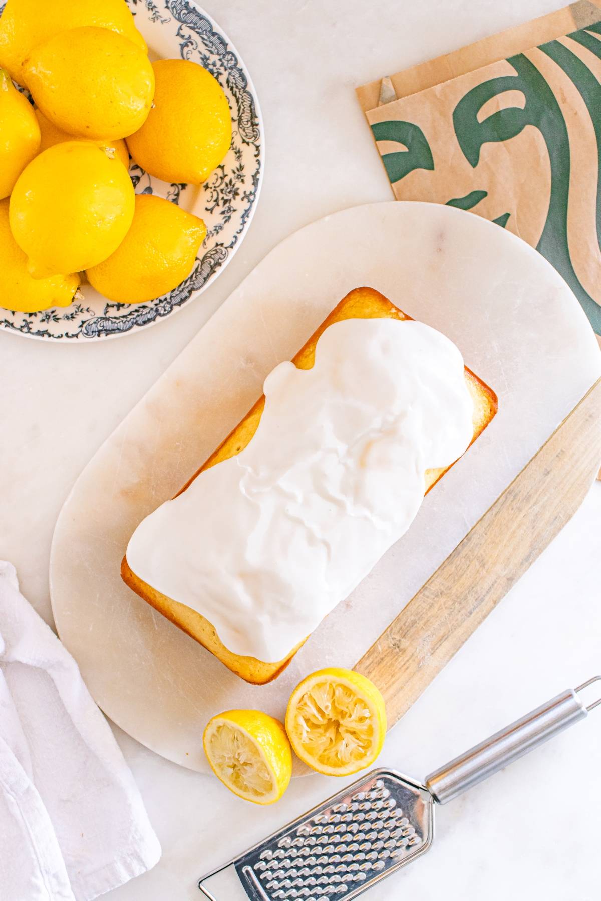 Iced lemon loaf on a board, with whole and halved lemons, a grater, and a patterned plate nearby.