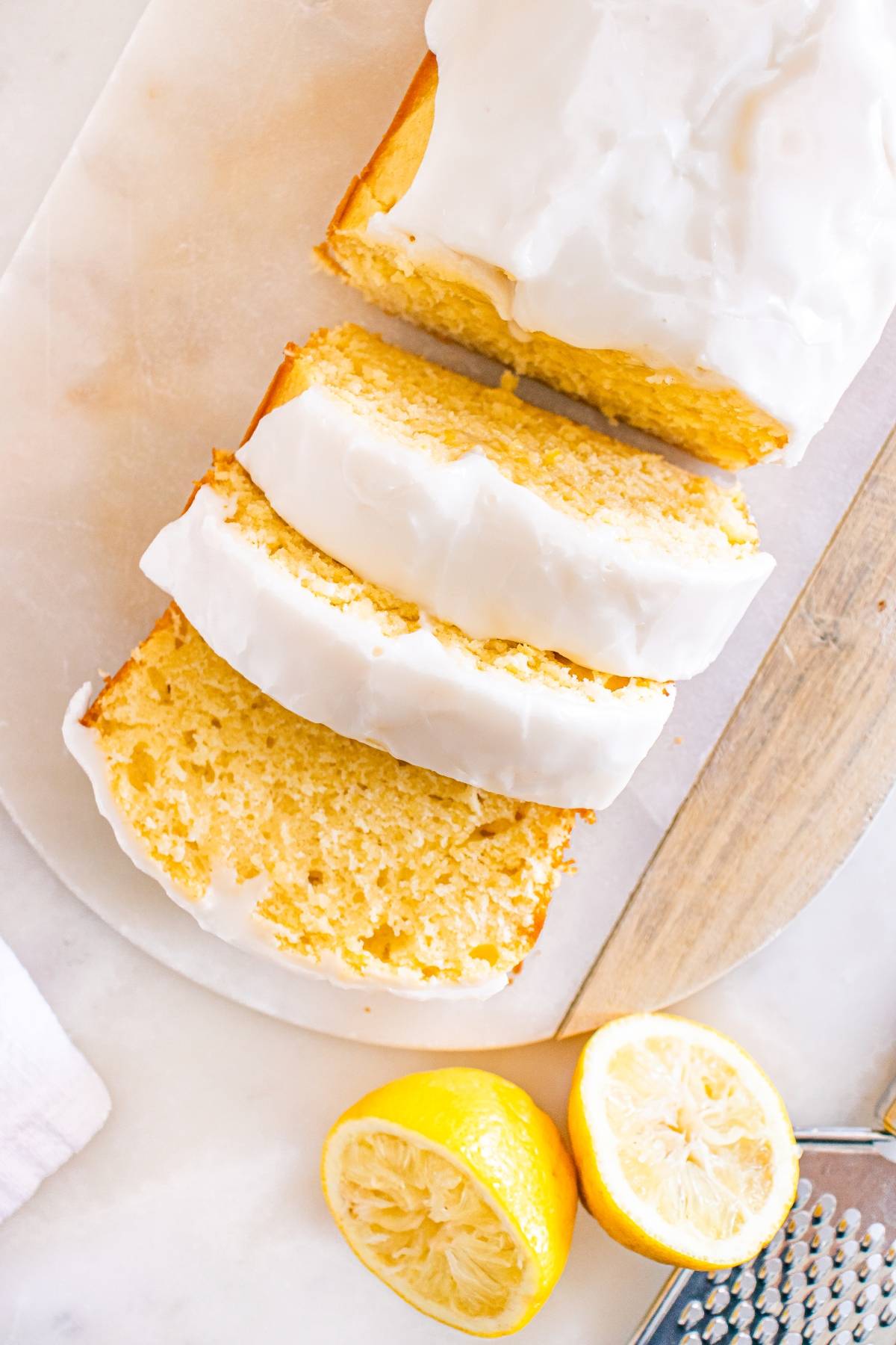 Sliced lemon loaf with white icing, next to a zested lemon and a grater on a marble surface.