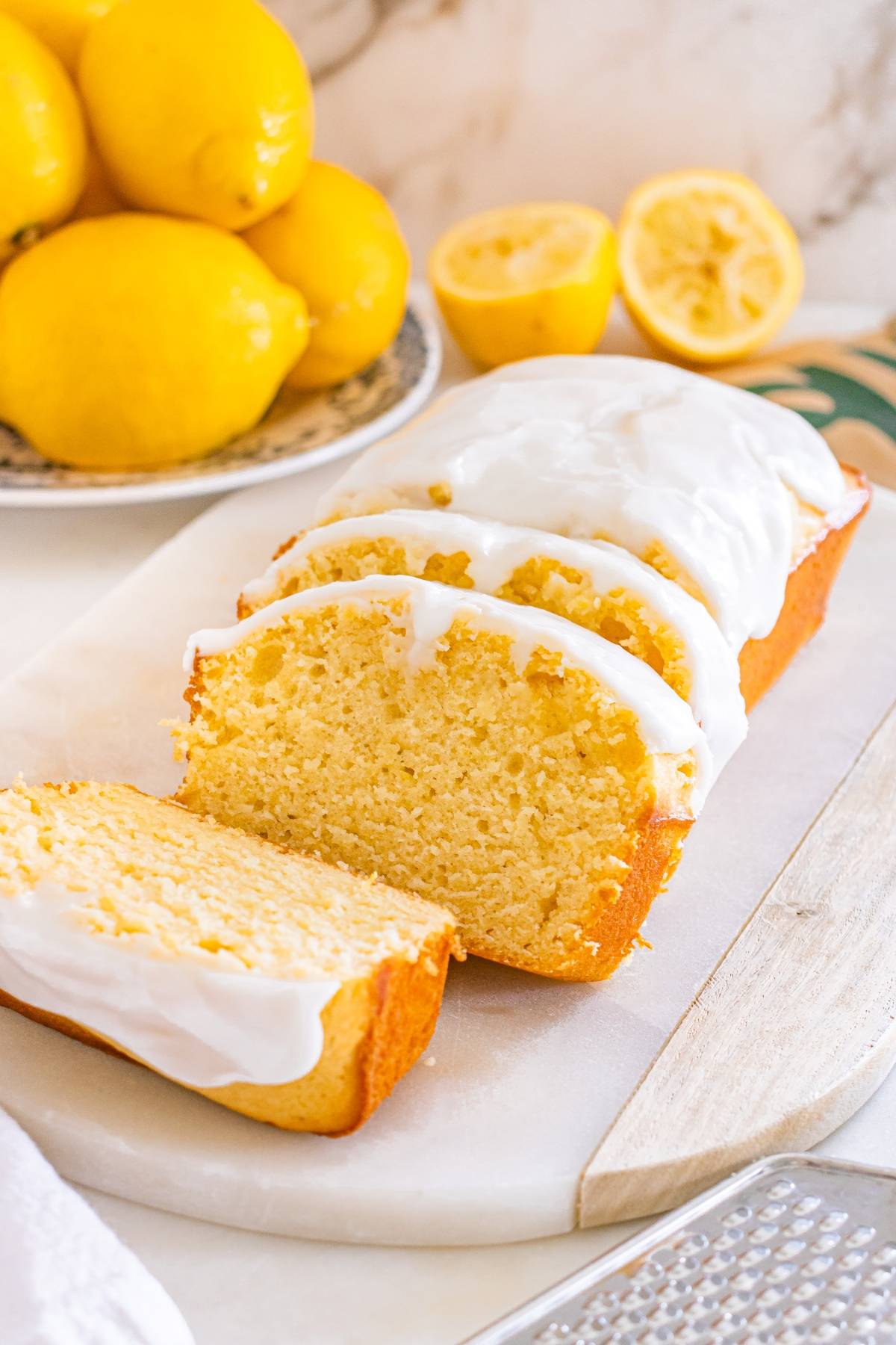 A loaf of lemon bread with white icing, partially sliced, sits on a marble board next to fresh lemons.