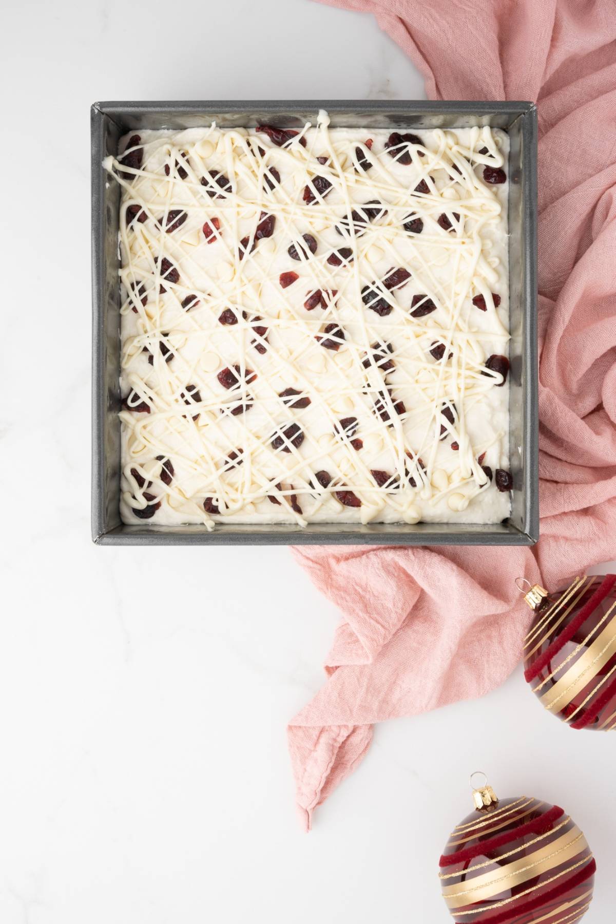 A square pan of white chocolate cranberry bars with cranberry pieces sits beside two ornaments and a pink cloth, capturing the homemade bliss of an easy cranberry bar for the holidays.