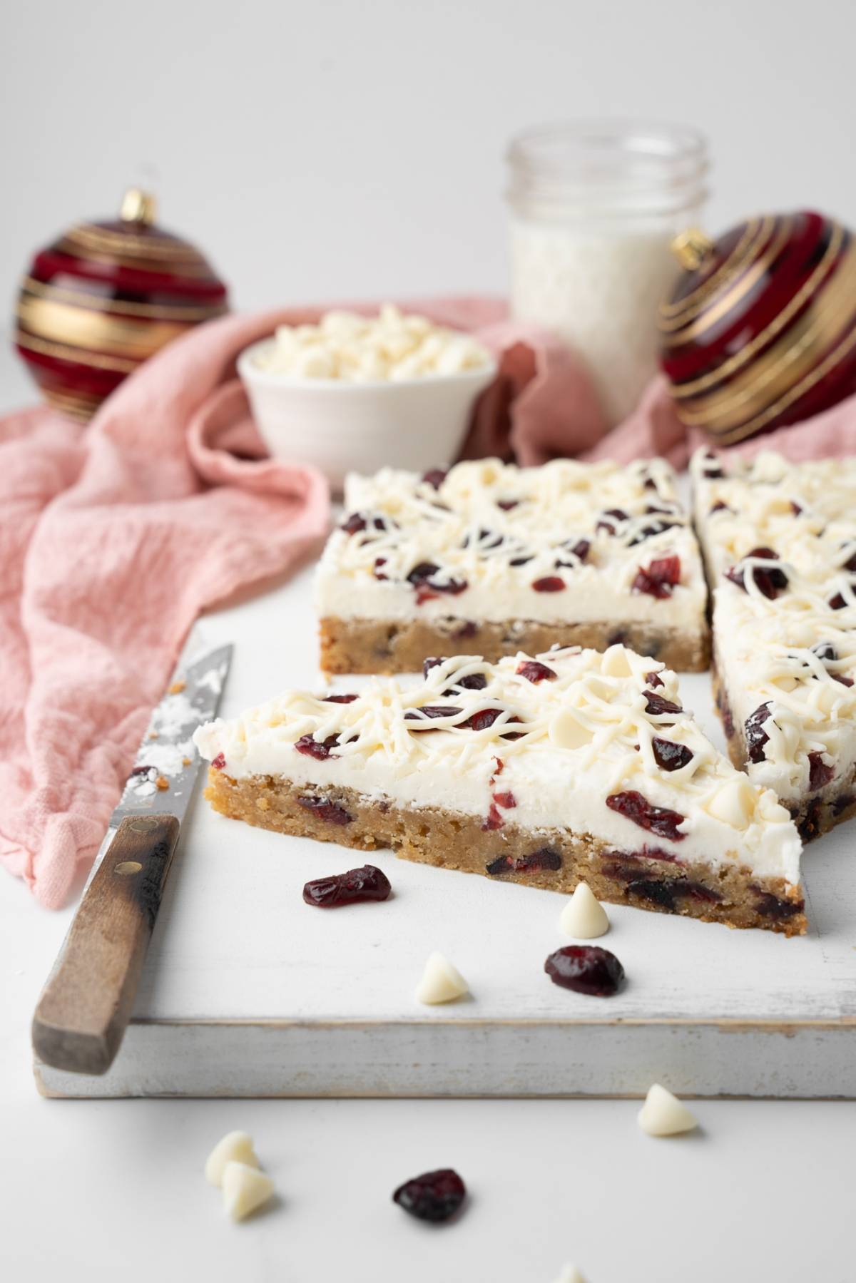 Three Starbucks copycat cranberry bliss bars sit on a white board with a knife, festive ornaments, and a pink cloth nearby.