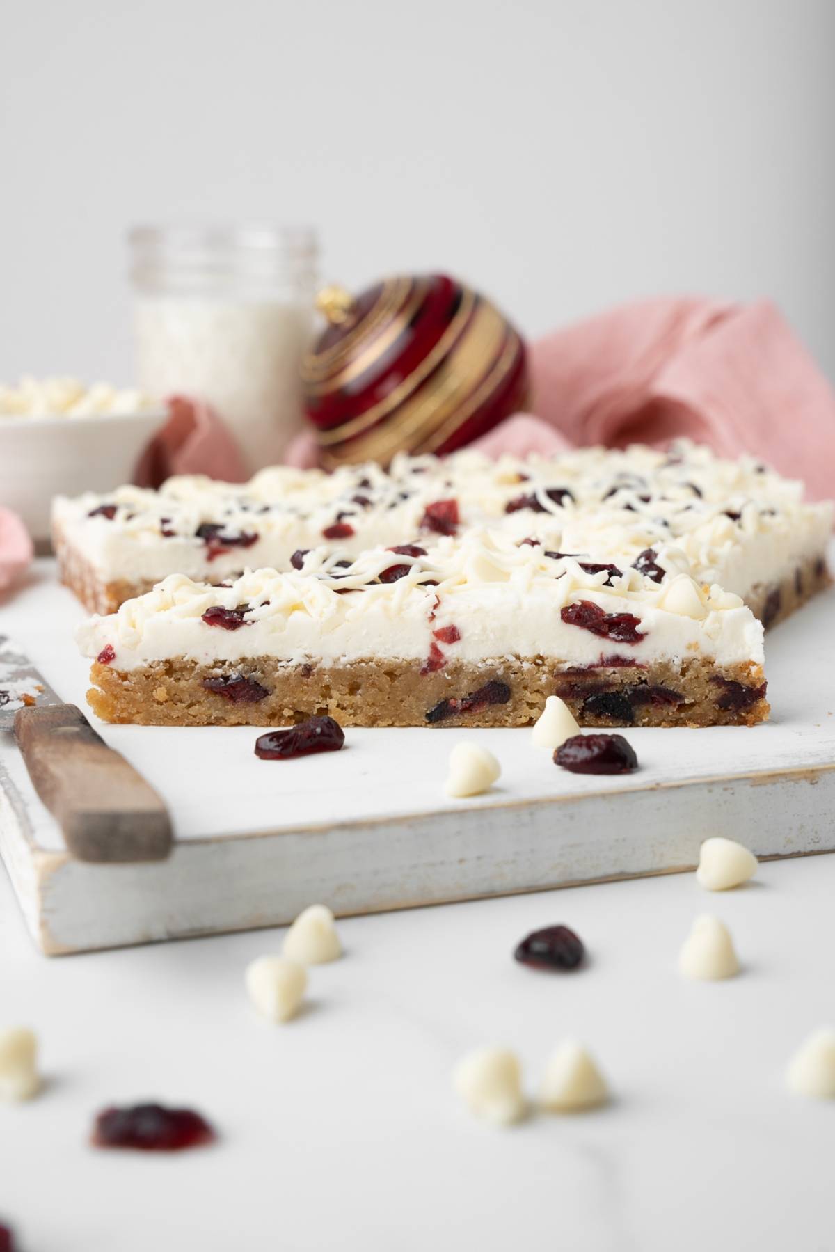 Frosted white chocolate cranberry bars with tart cranberries and creamy chips rest on a cutting board, surrounded by festive holiday decor—a classic, easy cranberry bar perfect for celebrations.