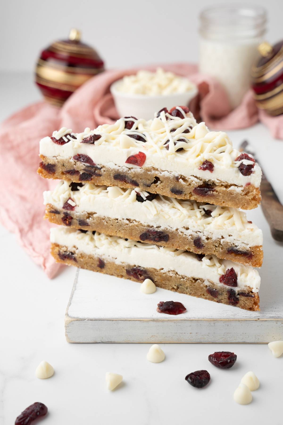 Three stacked white chocolate cranberry bars, reminiscent of easy cranberry bar treats, sit on a board with holiday ornaments in the background.
