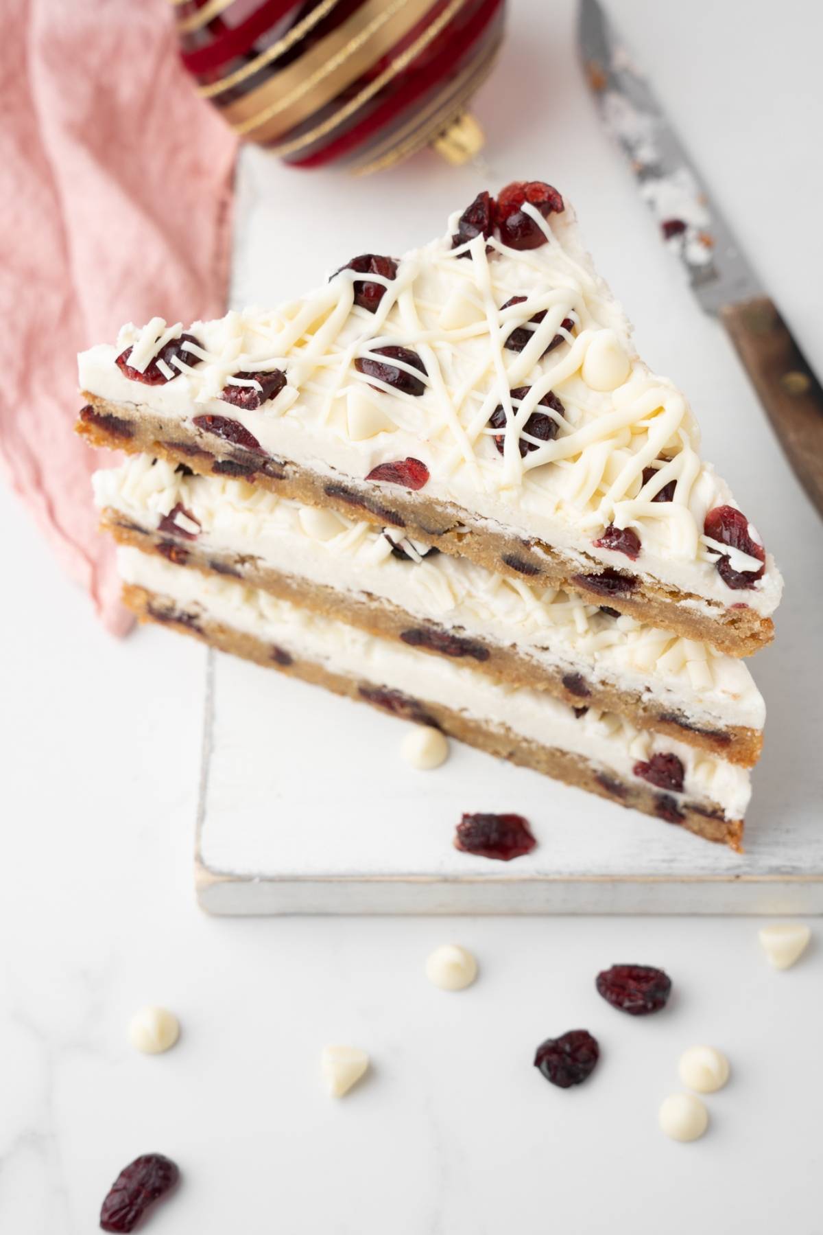 Three homemade bliss bars—white chocolate cranberry bars—are stacked on a board, with a knife and Christmas ornament nearby.