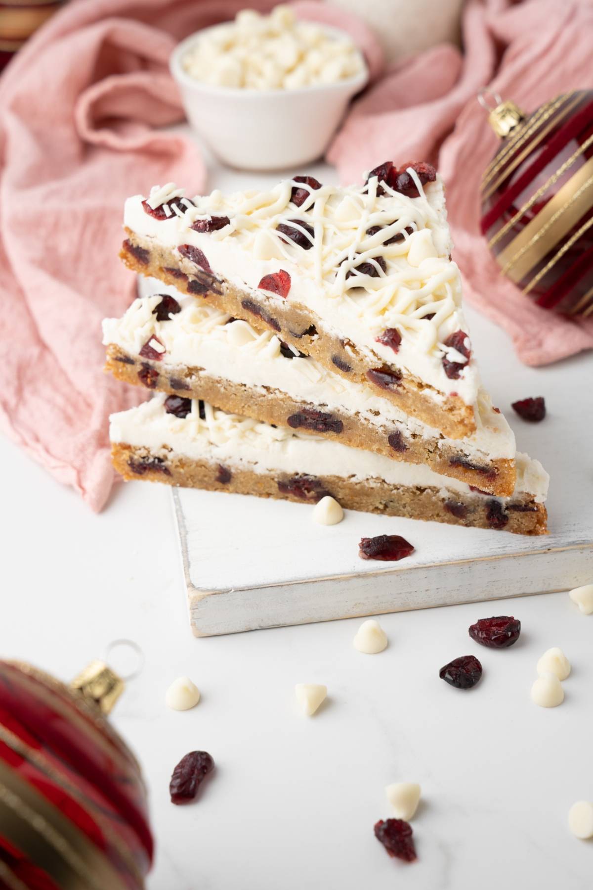 Three easy cranberry bars stacked on a white board, with ornaments and chocolate chips nearby, make these homemade bliss bars perfect for any festive occasion.