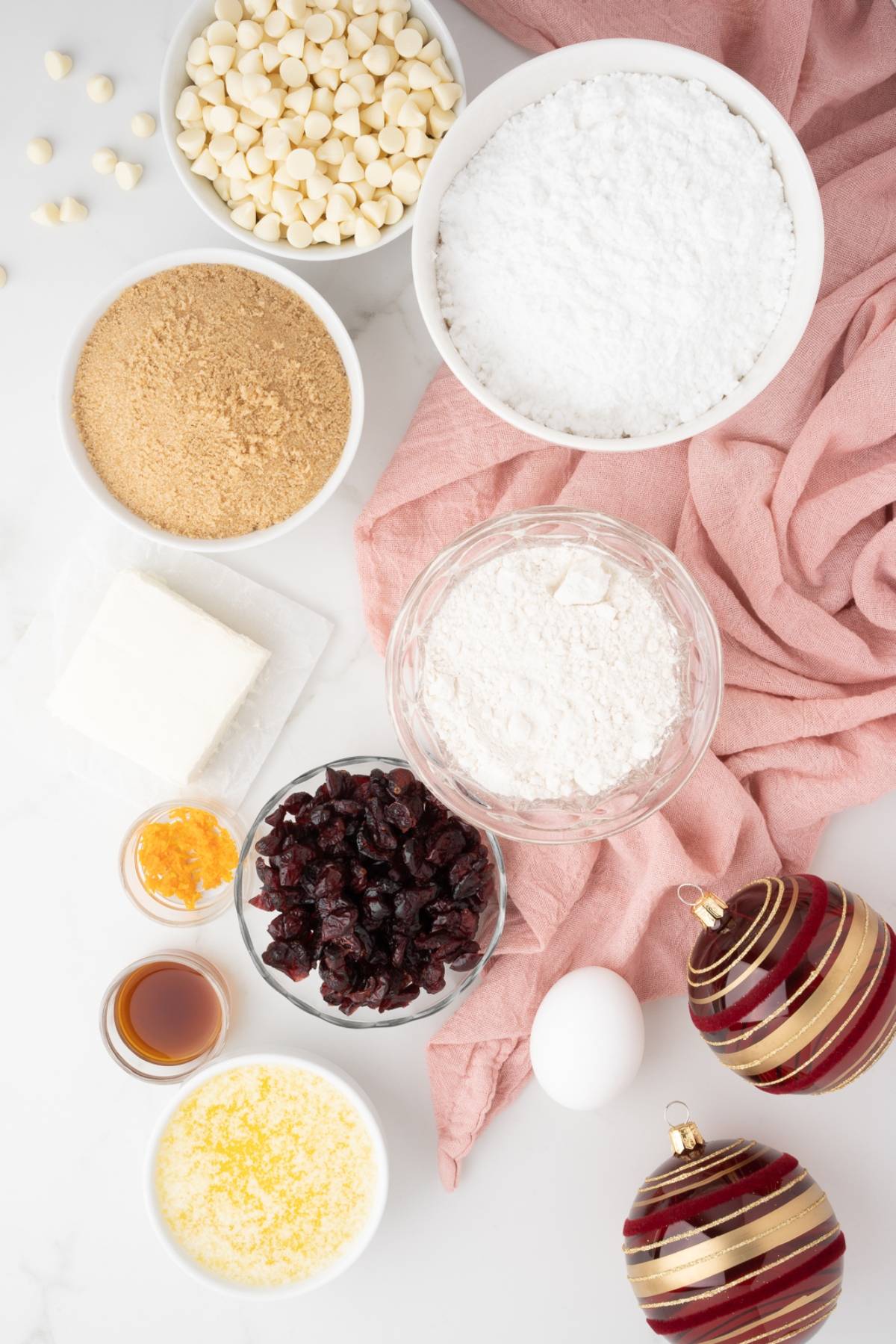 Baking ingredients in bowls with an egg, butter, orange zest, and holiday ornaments on a pink cloth—perfect for making easy cranberry bar or homemade bliss bars.