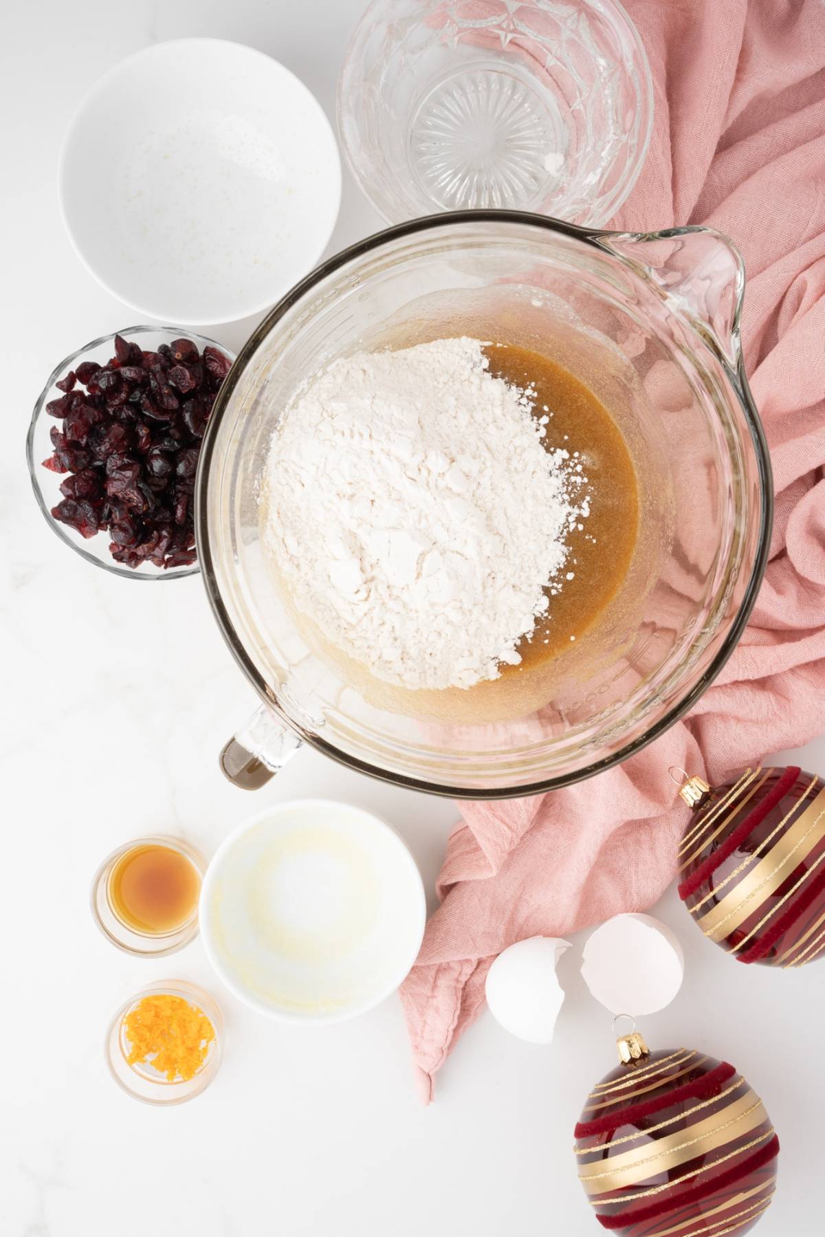 A mixing bowl with flour and wet ingredients, surrounded by cranberries, eggs, and holiday ornaments—perfect for creating easy cranberry bar recipes like Starbucks copycat cranberry bliss bars.