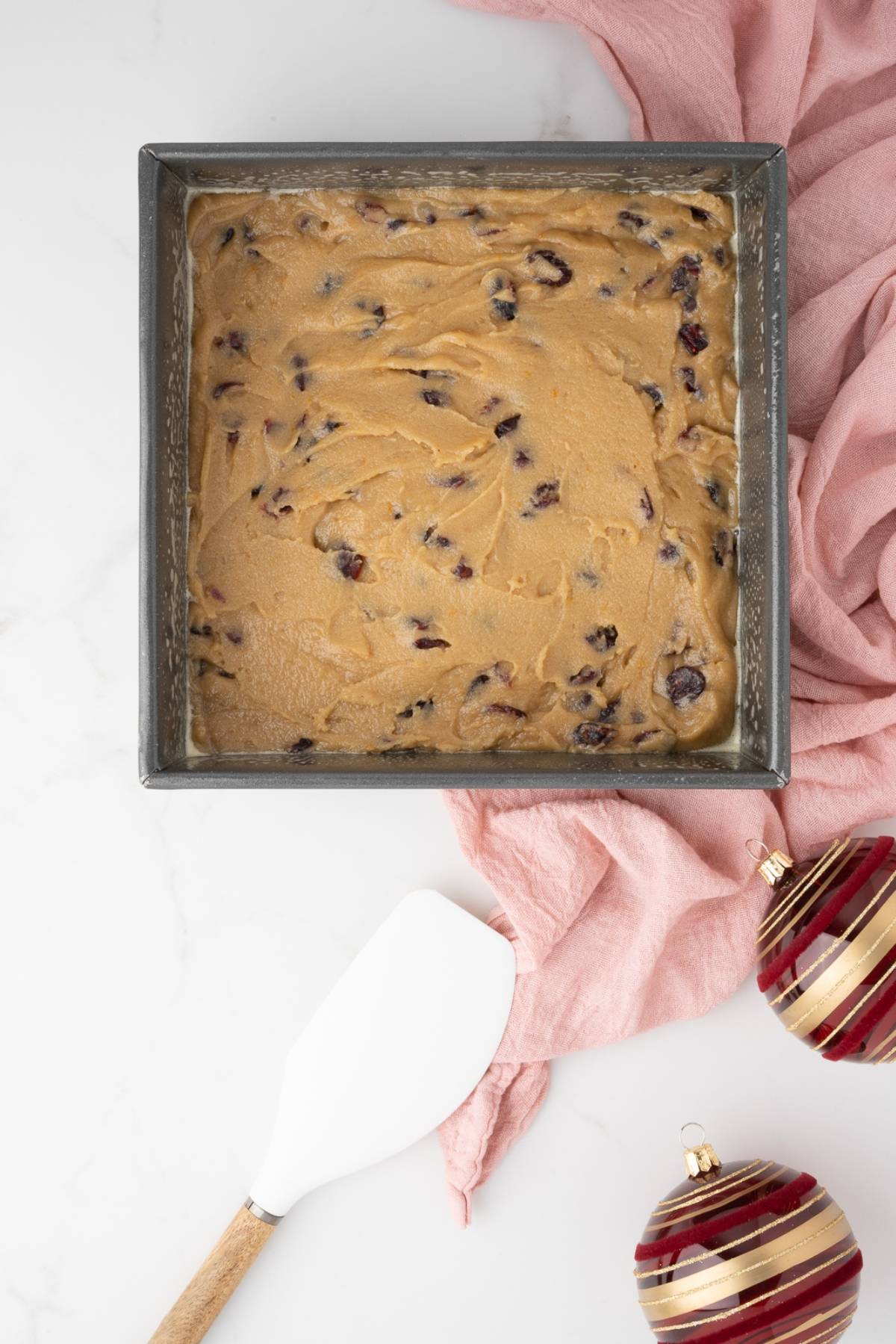 Square pan of cookie dough with chocolate chips, next to a spatula, pink cloth, and Christmas ornaments—perfect for baking white chocolate cranberry bars or your favorite Starbucks copycat cranberry bliss bars this holiday season.