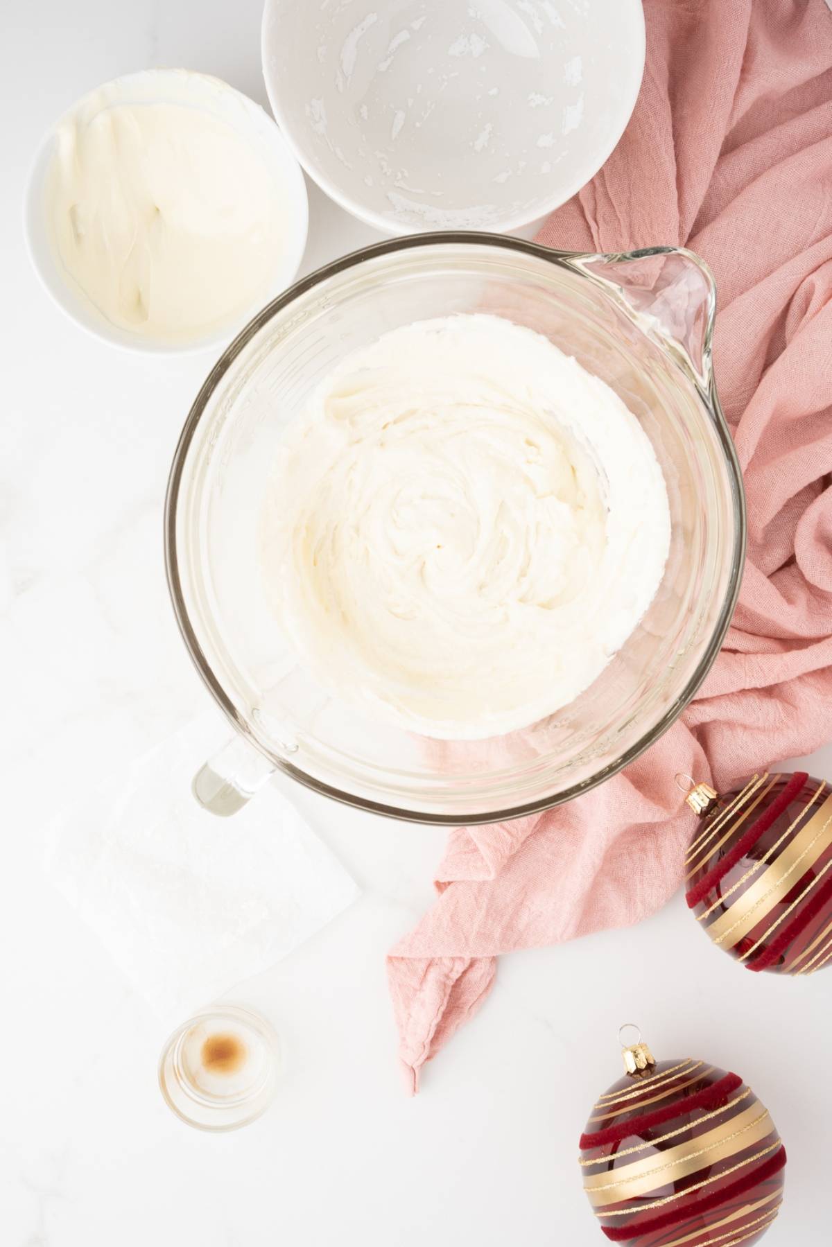 A bowl of whipped cream with bowls, a pink cloth, and Christmas ornaments on a white surface—perfect for pairing with homemade bliss bars or your favorite easy cranberry bar this holiday season.