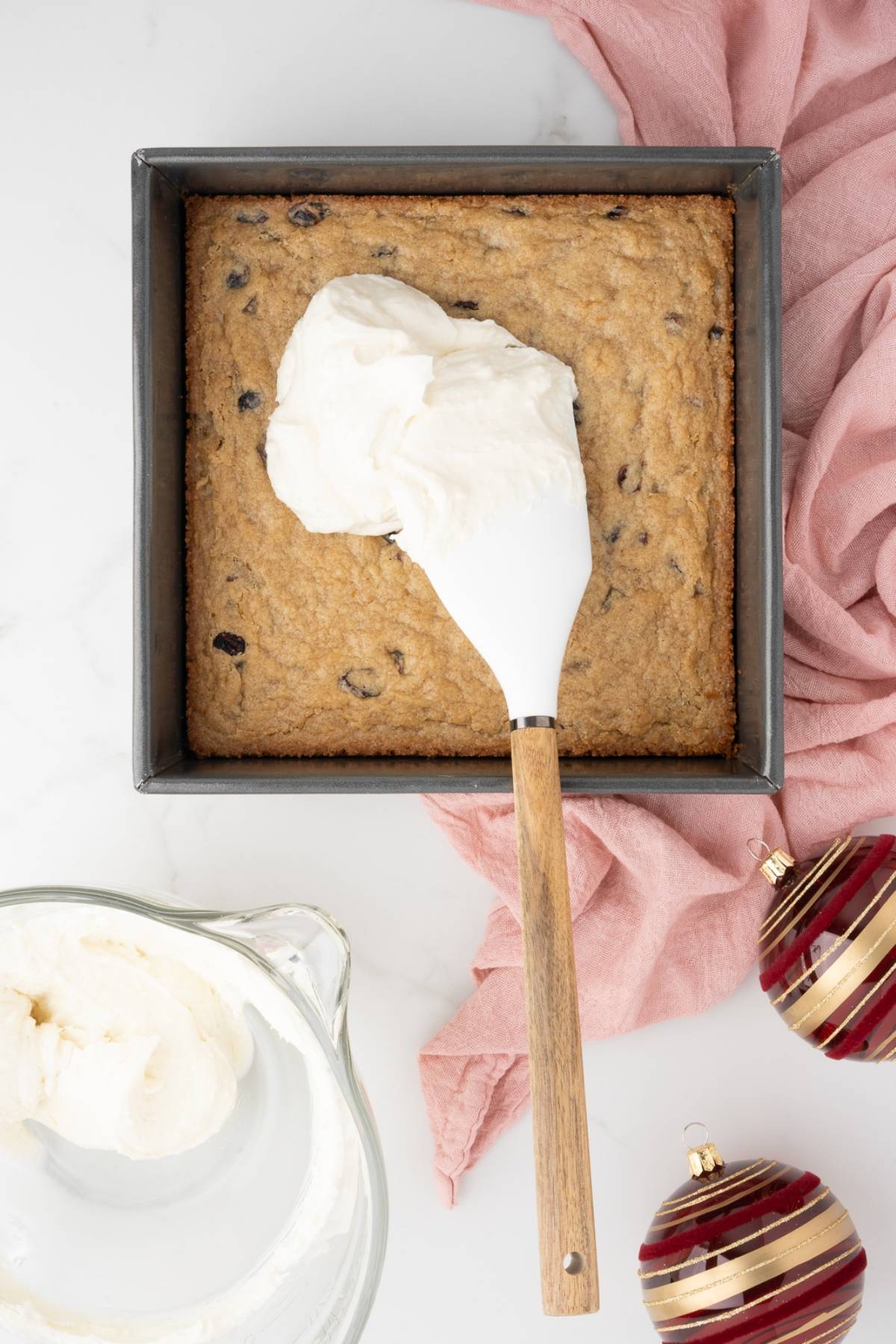 A spatula spreads frosting on a square cake in a pan, resembling easy cranberry bars, with ornaments and a pink cloth nearby.
