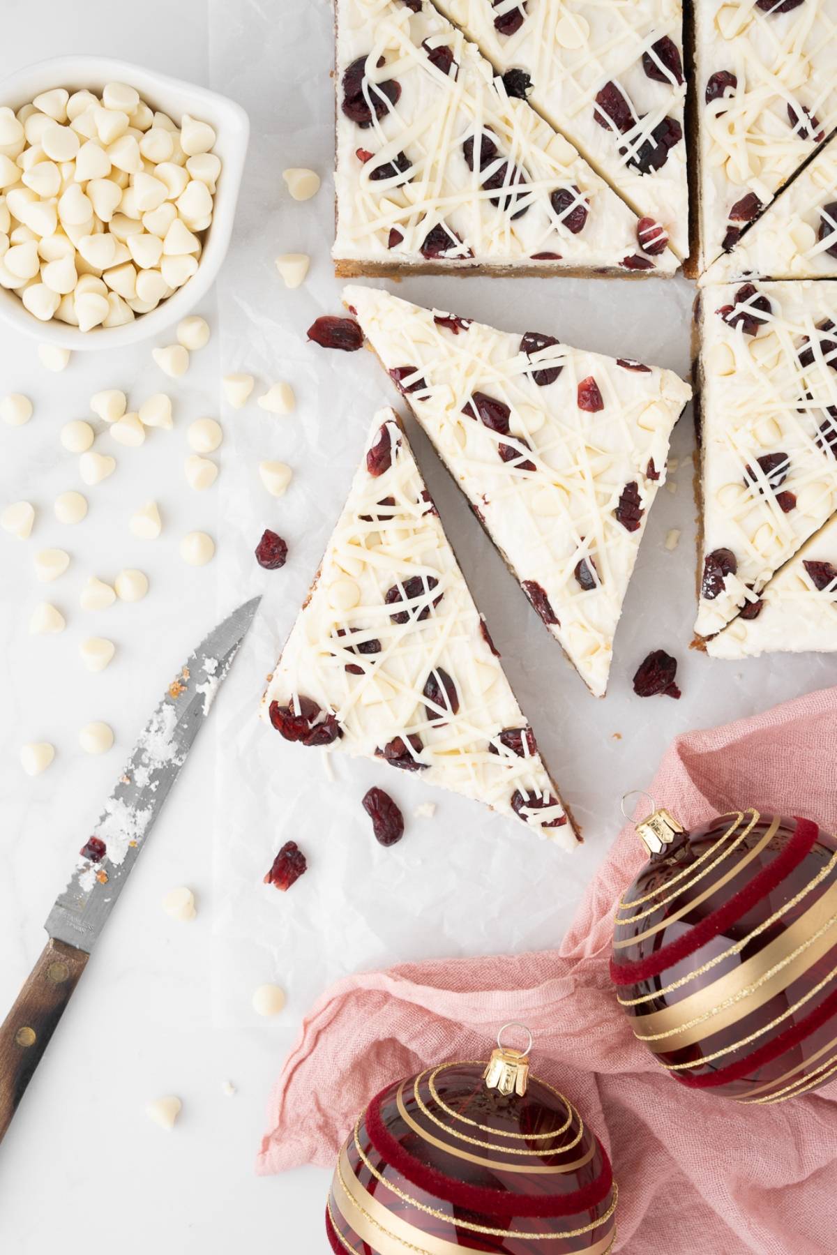 Sliced Starbucks copycat cranberry bliss bars with white chocolate drizzle, a knife, white chocolate chips, and Christmas ornaments.