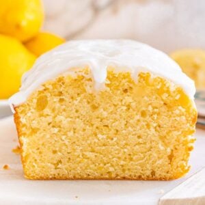 A Starbucks copycat lemon loaf with white icing is sliced on a white surface, with fresh lemons in the background.