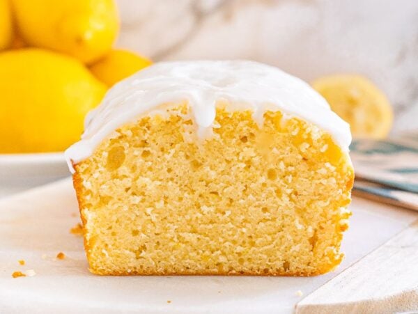 A Starbucks copycat lemon loaf with white icing is sliced on a white surface, with fresh lemons in the background.
