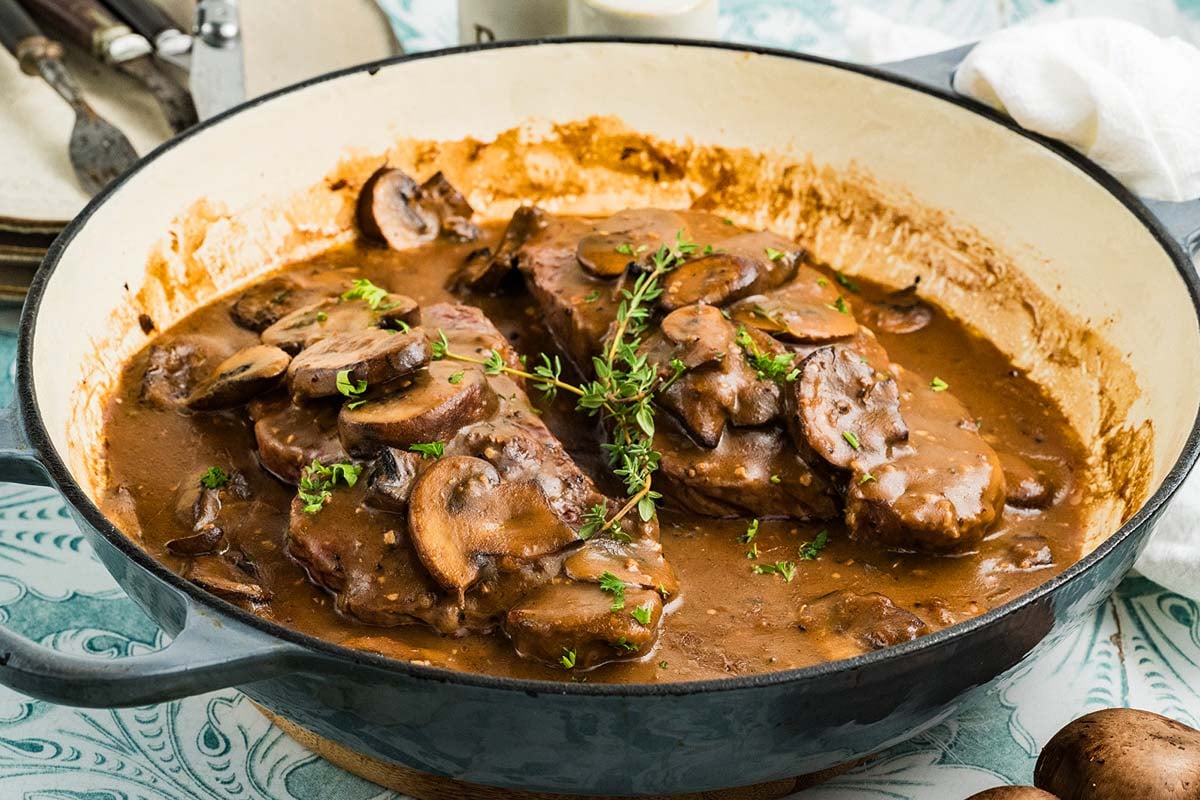 Steak marsala in a rich mushroom gravy garnished with herbs and served in a large skillet.