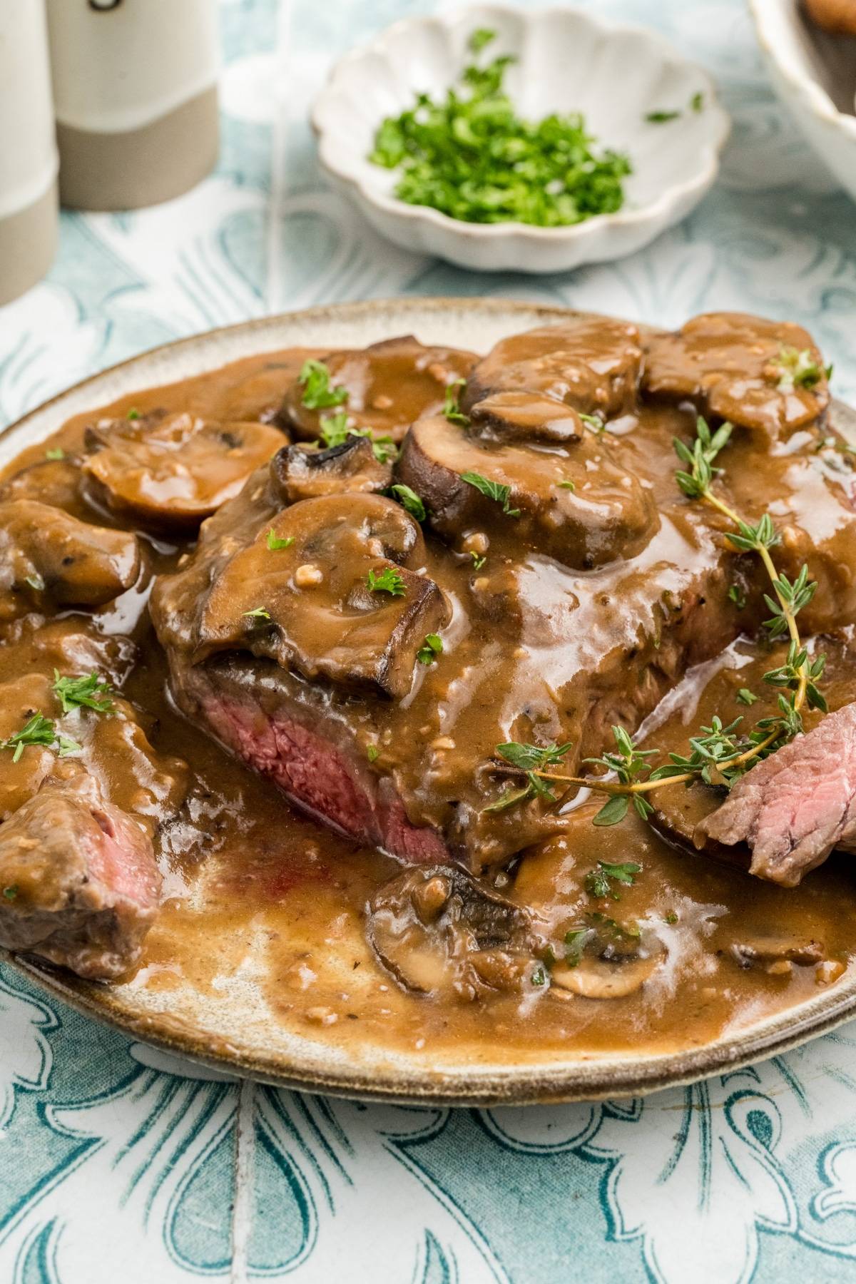 Sliced steak topped with mushroom gravy and fresh herbs on a plate, with a garnish dish in the background.