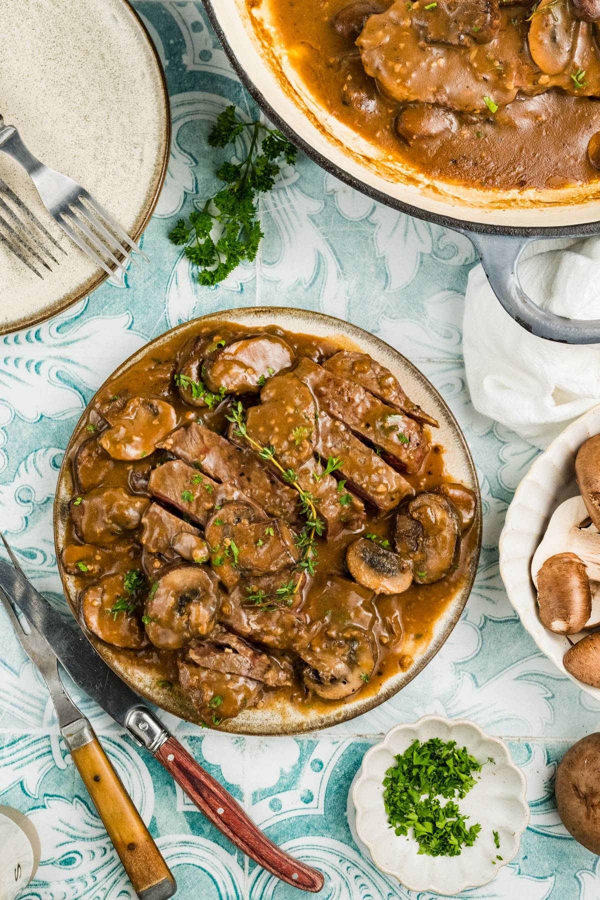 Plate of beef marsala with brown mushroom gravy, garnished with herbs, on a blue-patterned table with utensils and ingredients.