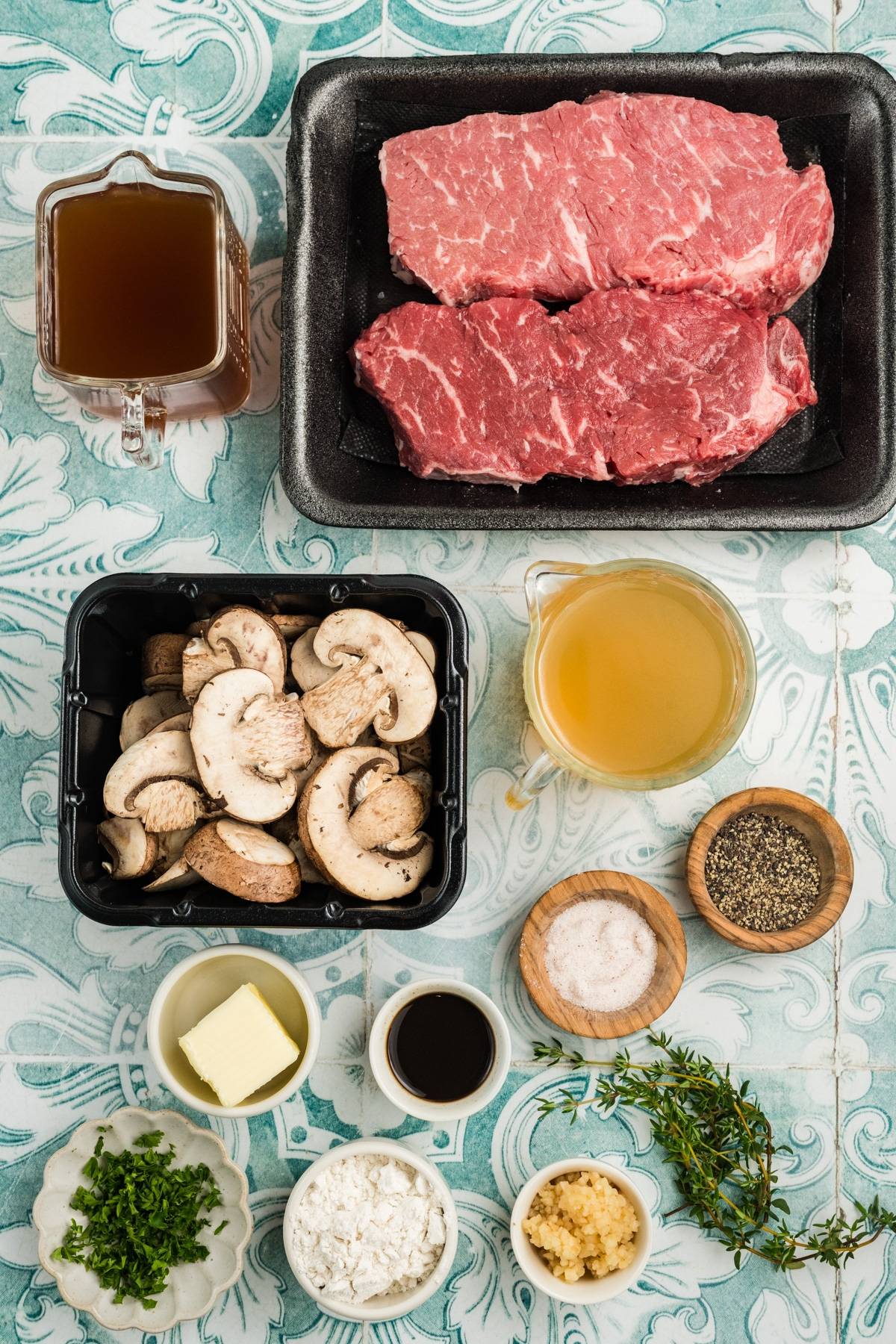 Raw beef, sliced mushrooms, broth, seasonings, herbs, butter, garlic, and sauce on a patterned surface.