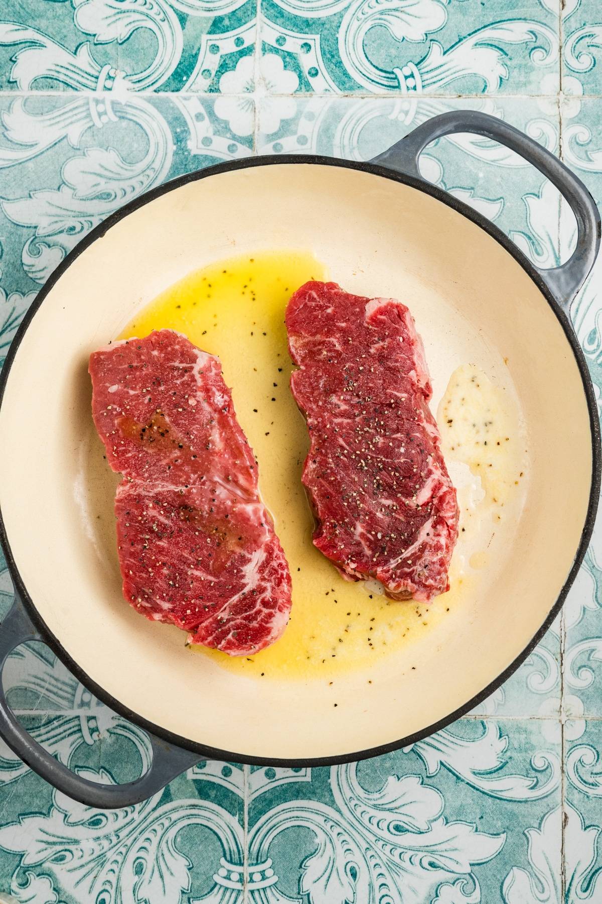 Two raw steaks seasoned with pepper in a white pan with oil, on a blue patterned tile surface.