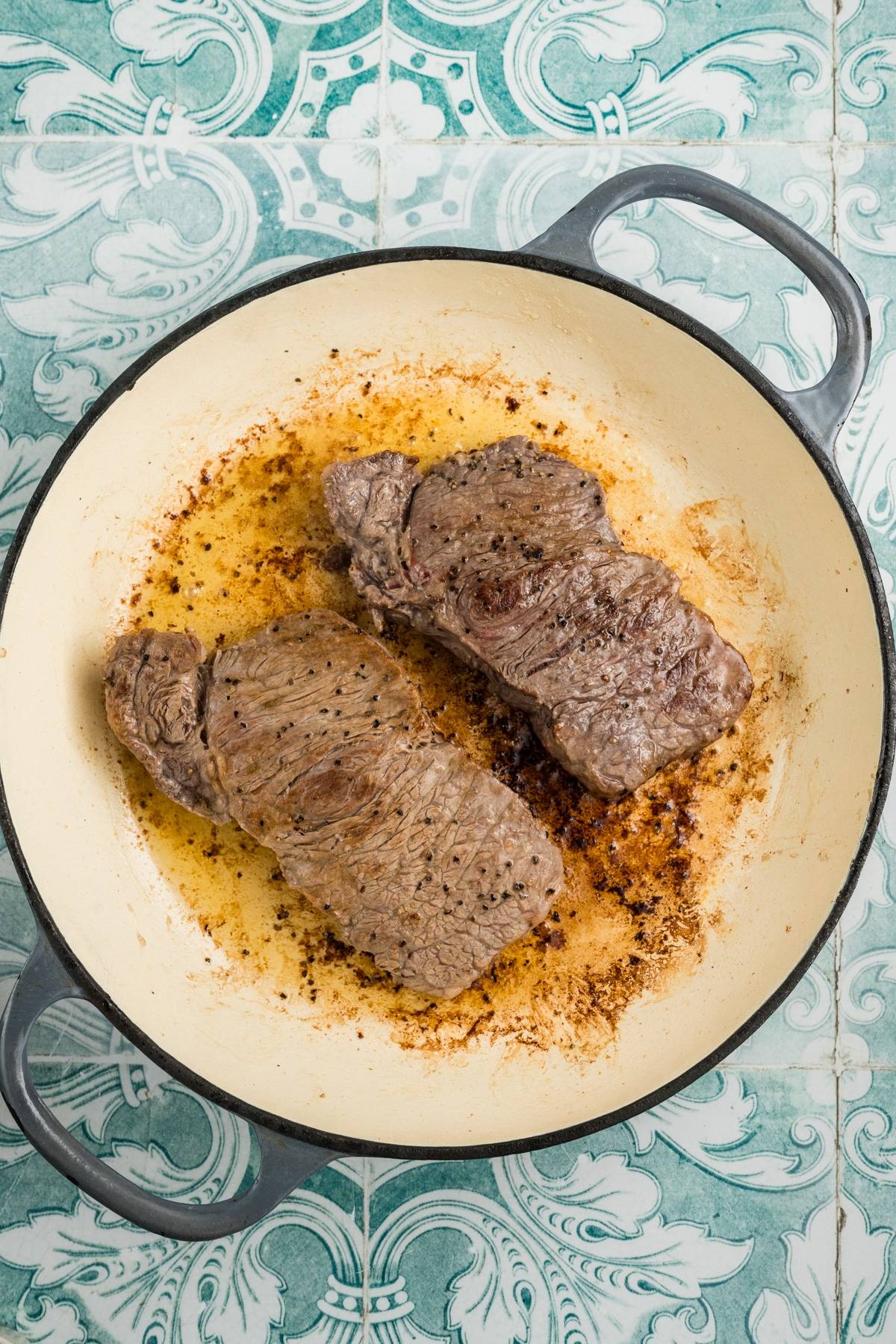 Two seared steaks cooking in a round, white pan with handles on a patterned green tile surface.